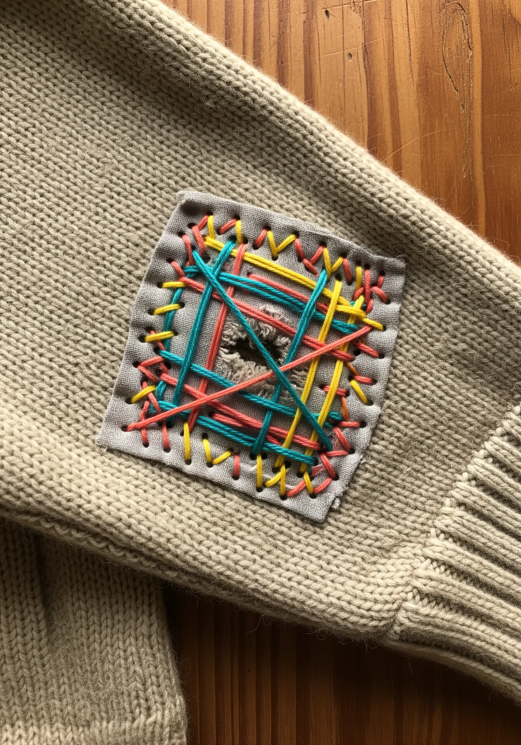 A woven mending patch with colorful threads on the elbow of a beige knit sweater.