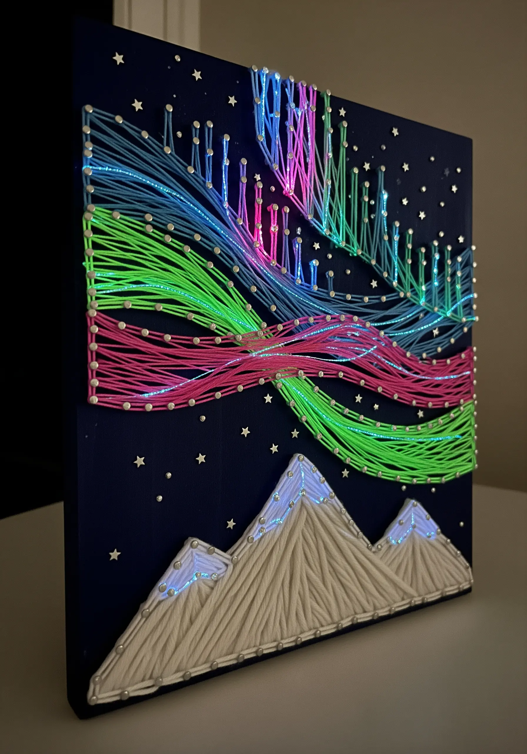 String art of the northern lights in neon colors above snowy mountains, with small lights.