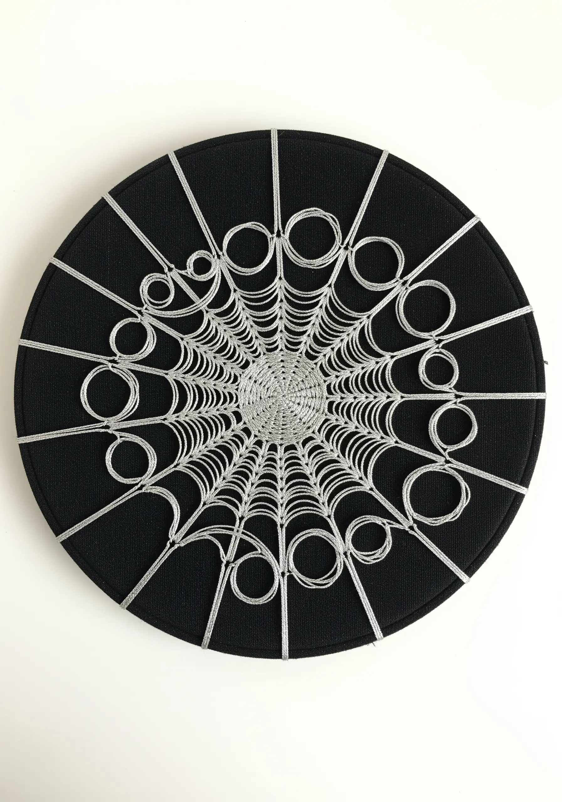 A sculptural spider web made with silver cord on a black circular canvas