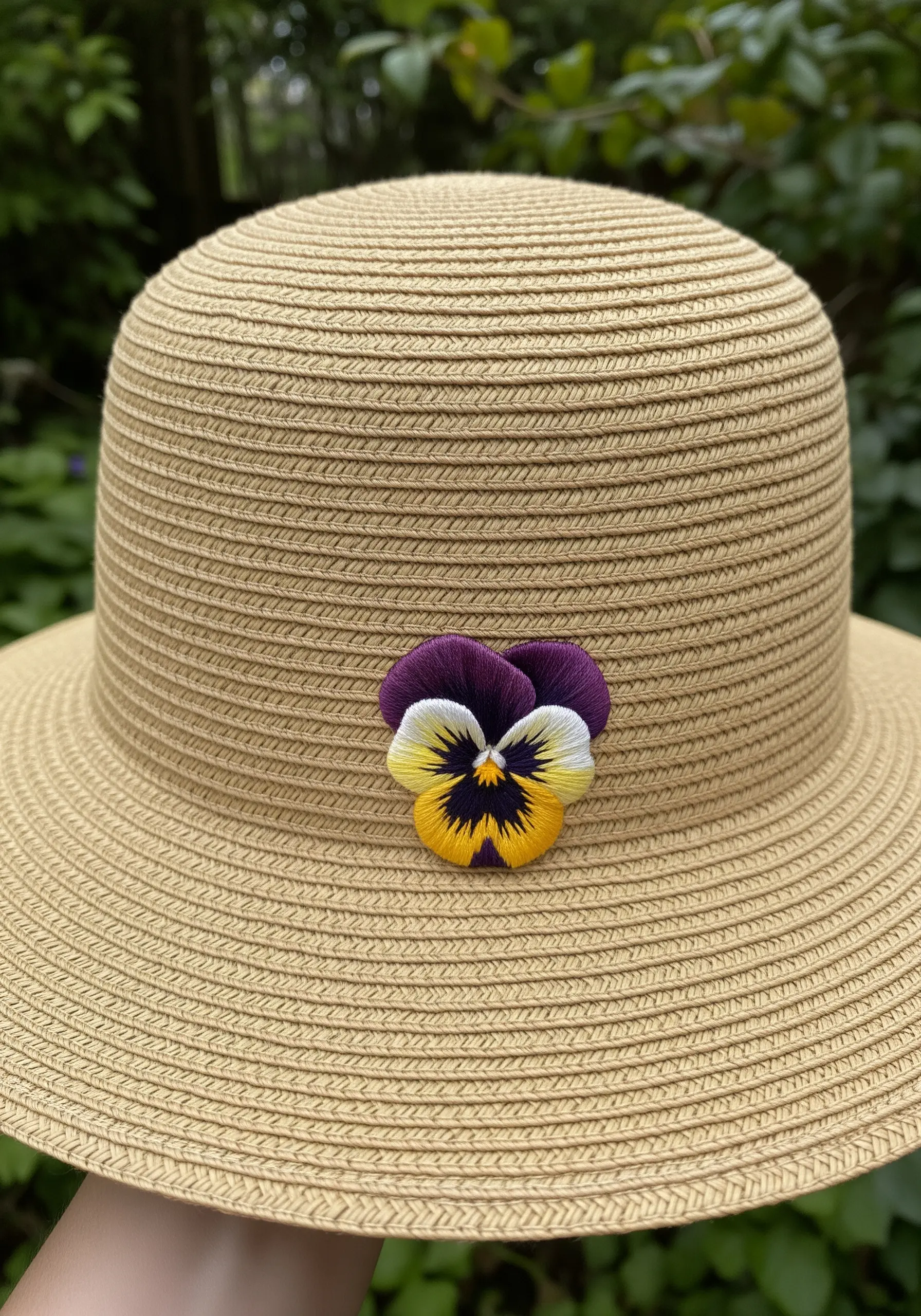 A detailed, thread-painted pansy with purple, yellow, and white petals on a straw hat.