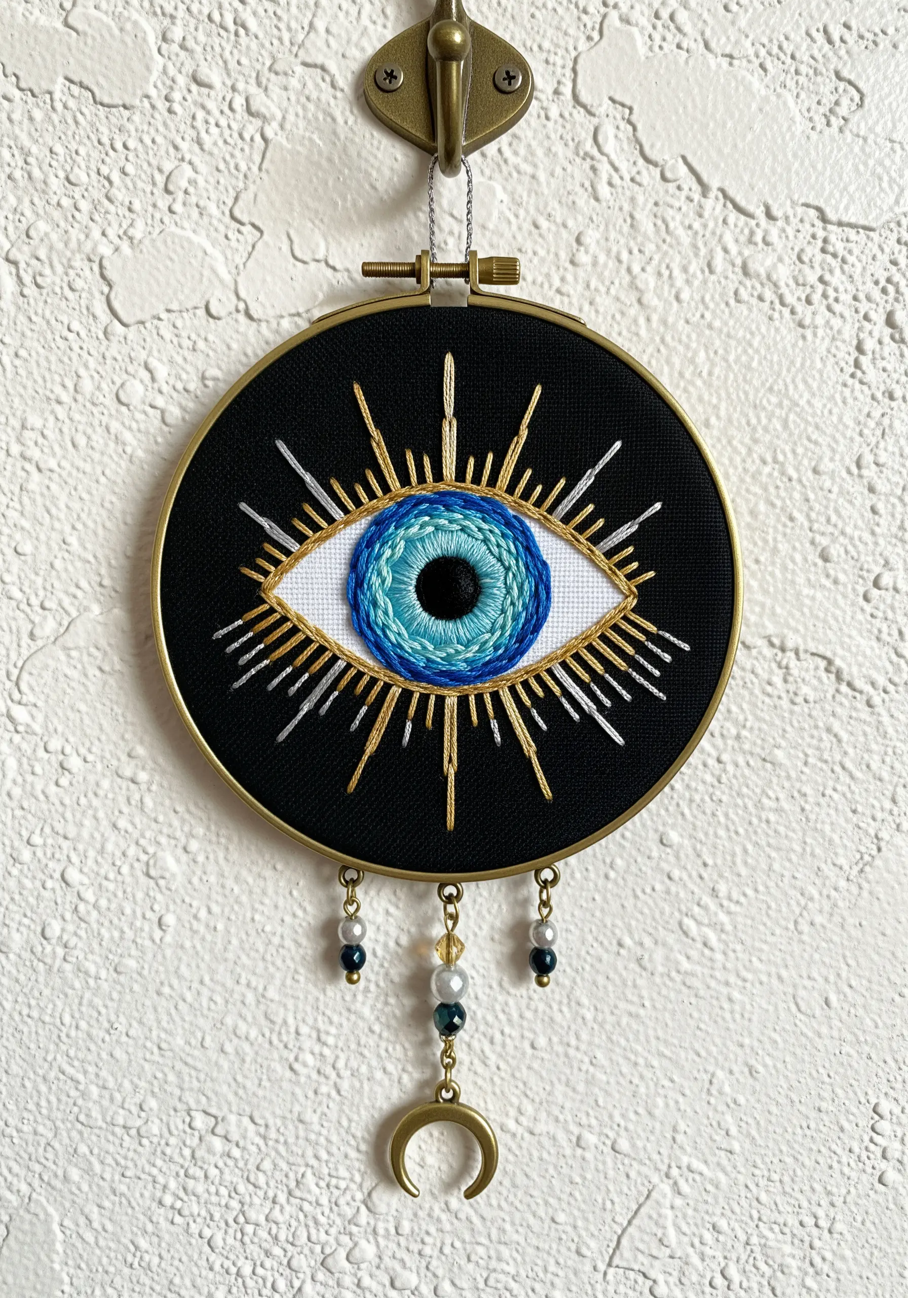 Embroidered evil eye talisman on black fabric with gold rays and bead dangles.