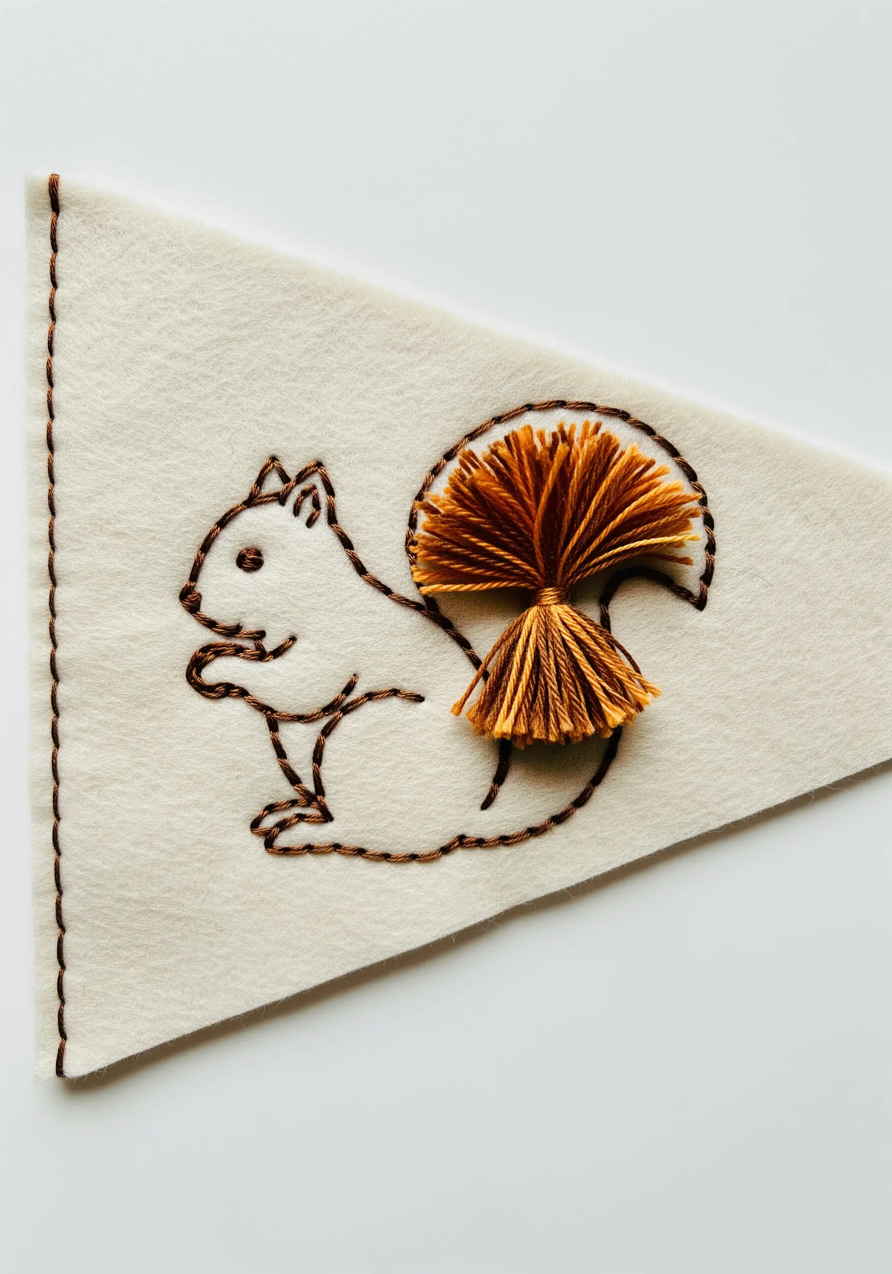 A line-art squirrel with a fluffy, three-dimensional tassel for its tail on a felt pennant.