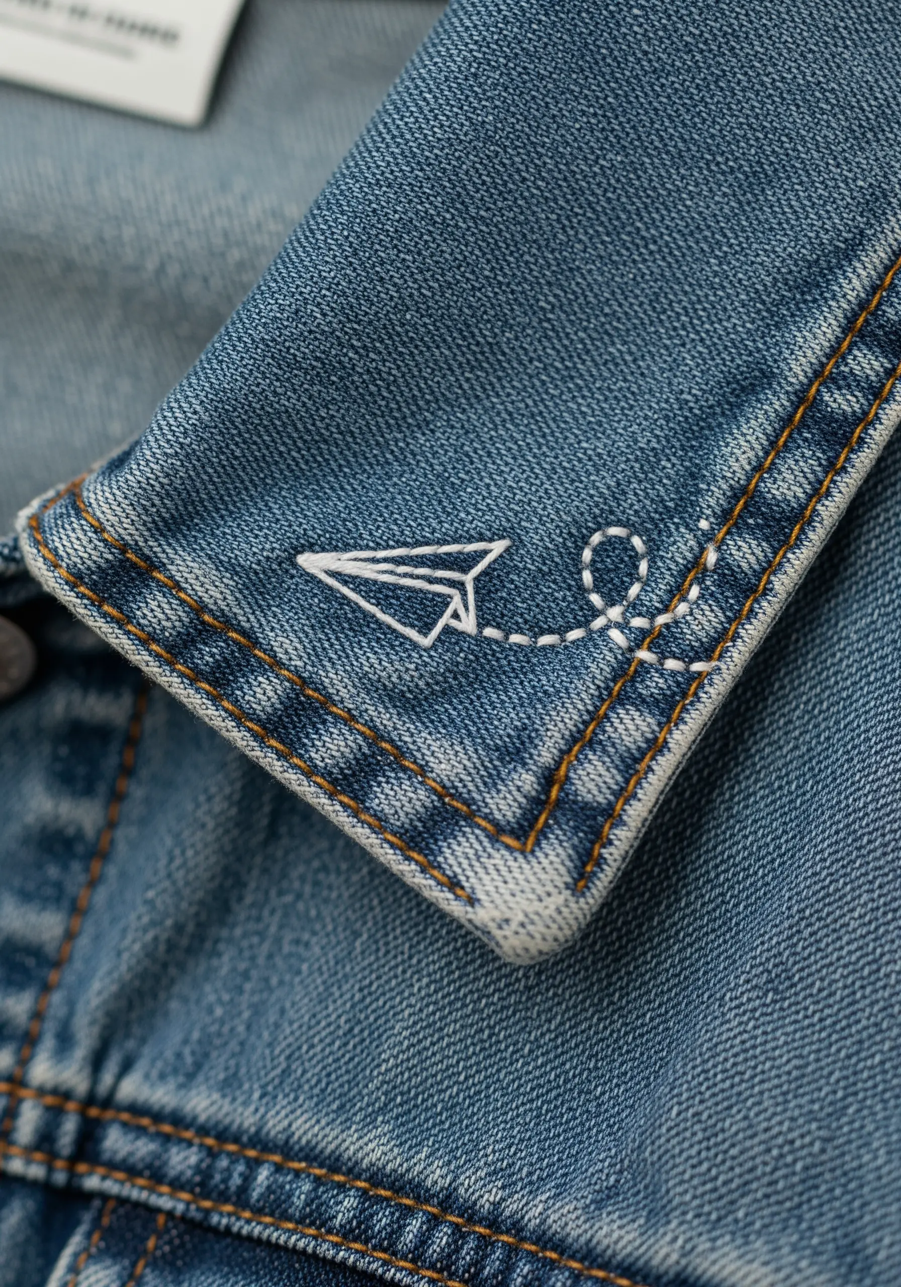 A small white paper airplane embroidered on the collar of a denim jacket.