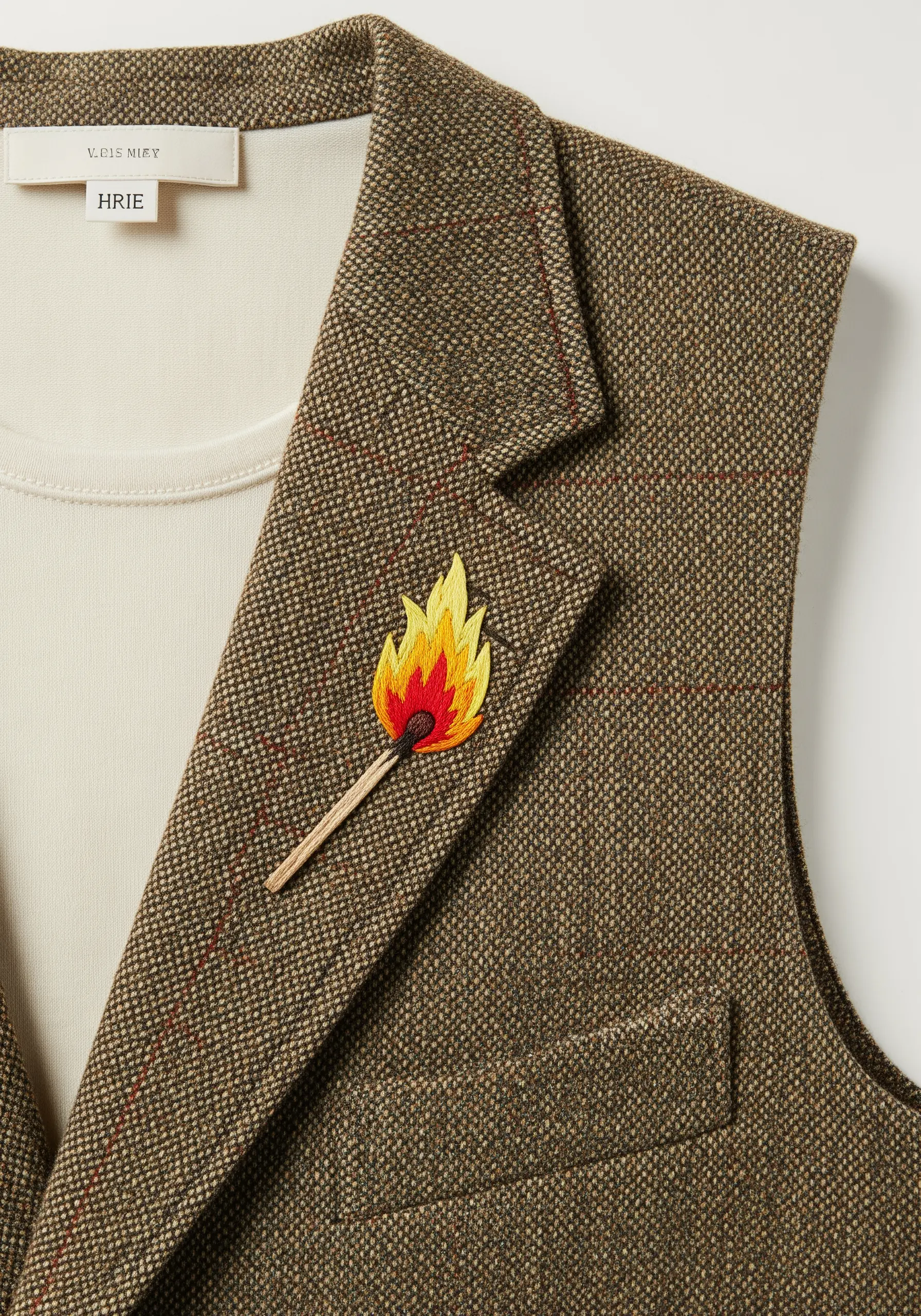 A small, colorful embroidery of a lit matchstick on the lapel of a brown tweed vest.