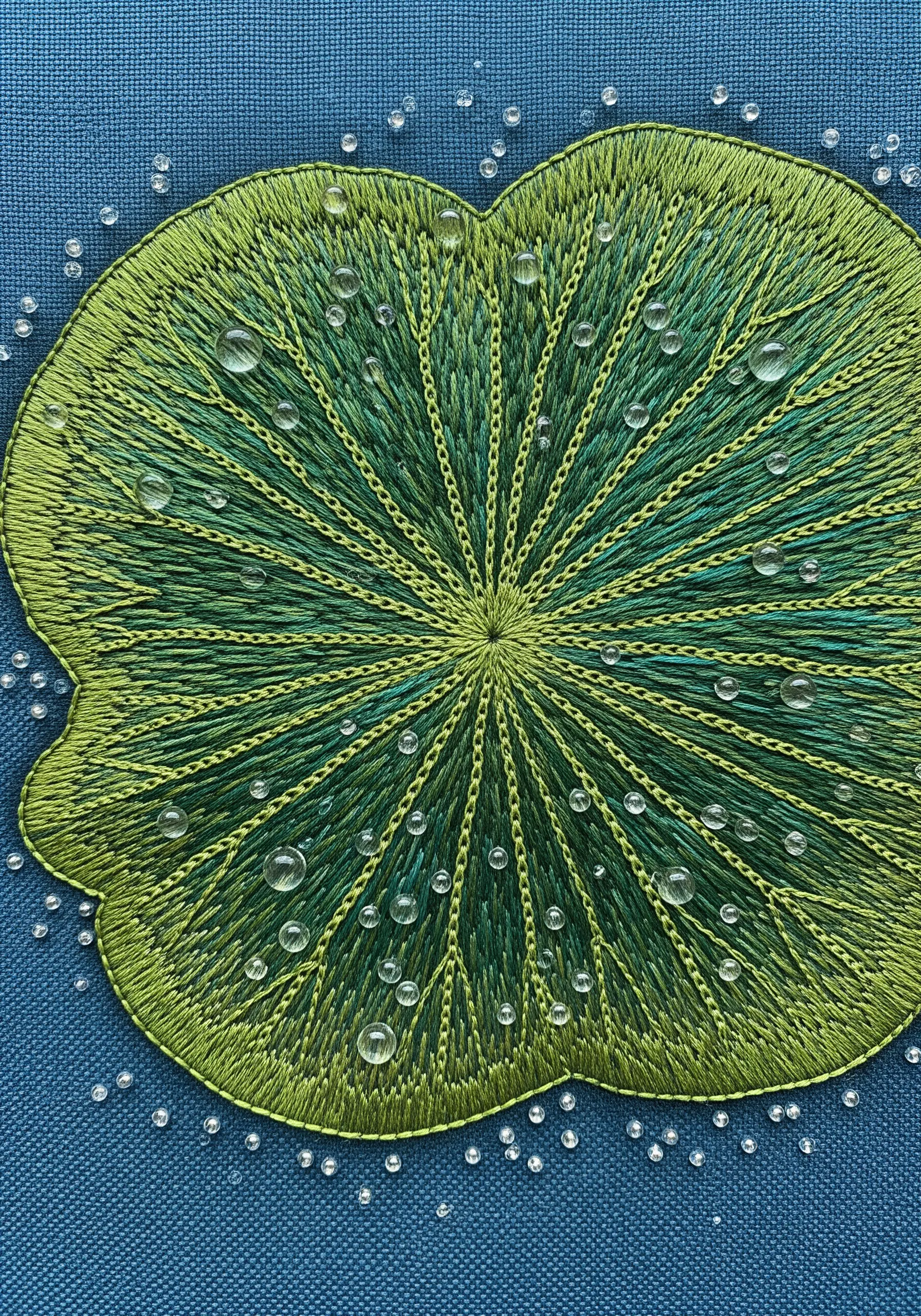 A green lily pad embroidered with radiating stitches and decorated with clear seed beads.