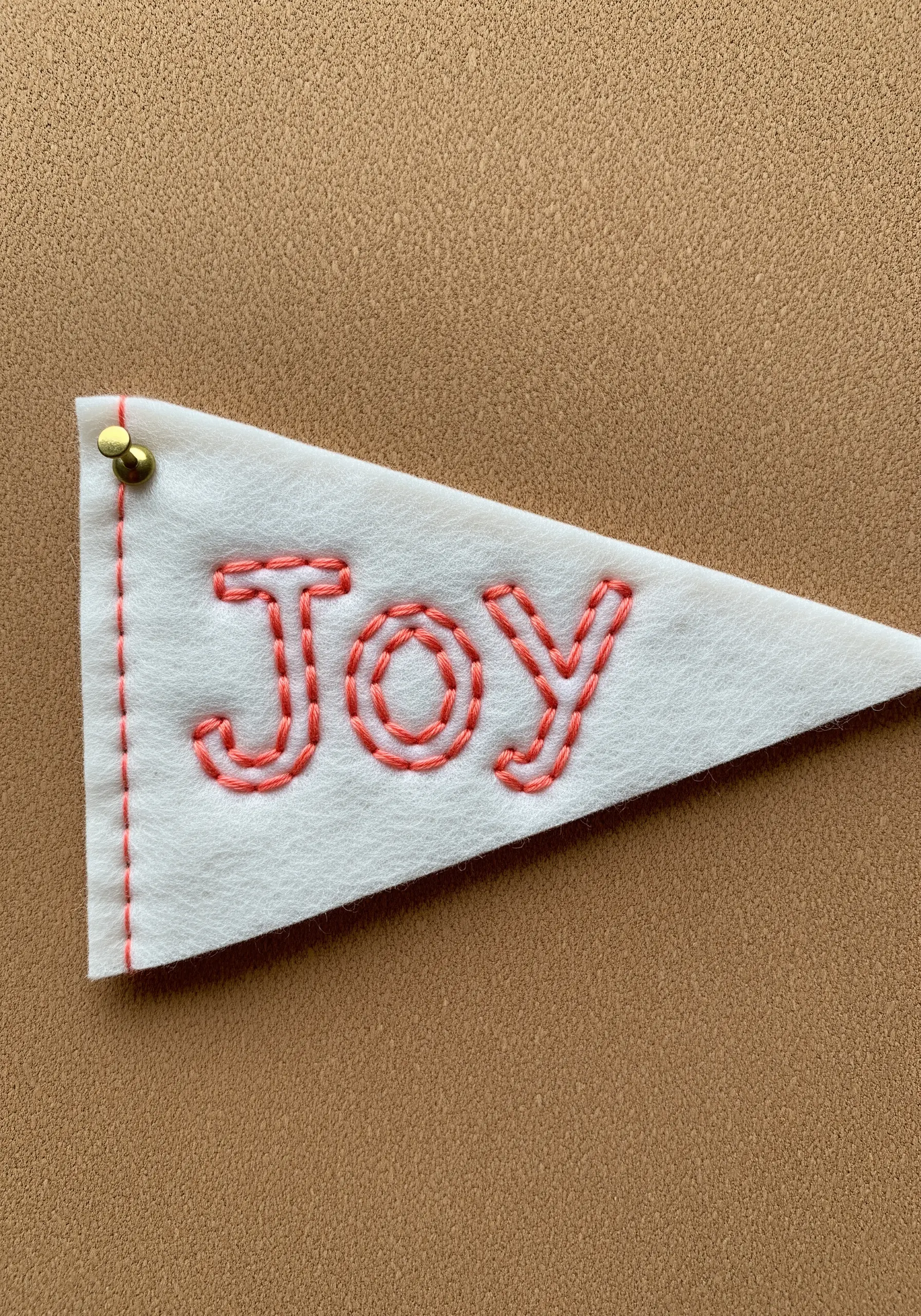 The word 'Joy' embroidered in coral whipped backstitch on a small white felt pennant.