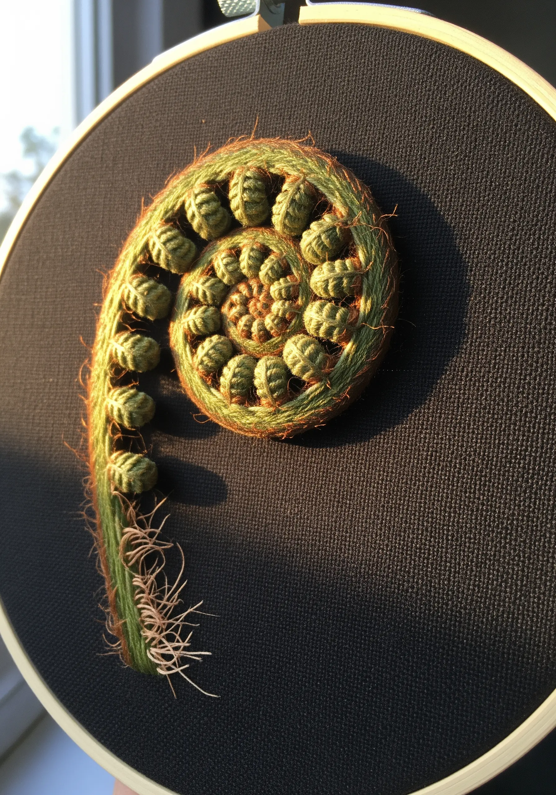 A dimensional embroidery of a curled fiddlehead fern using padded satin stitch and fuzzy yarn.