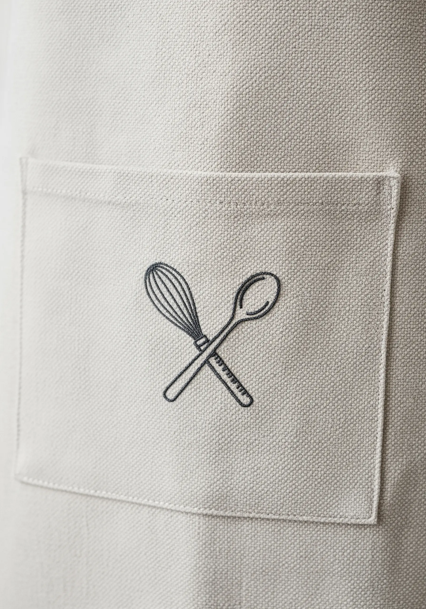 Line art embroidery of a crossed whisk and spoon on an apron pocket.