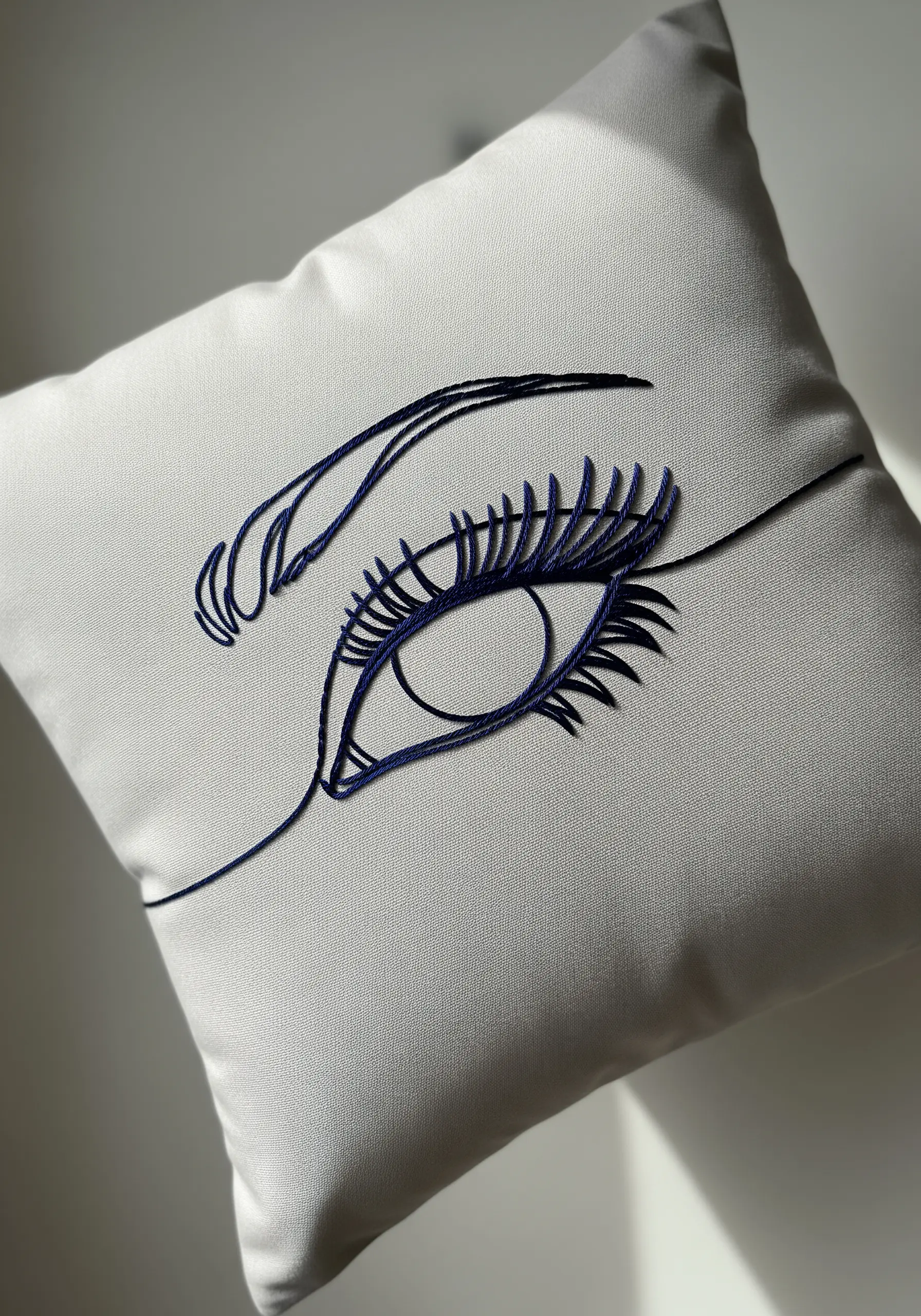 Close-up of an embroidered eye in dark blue thread on a light grey cushion.