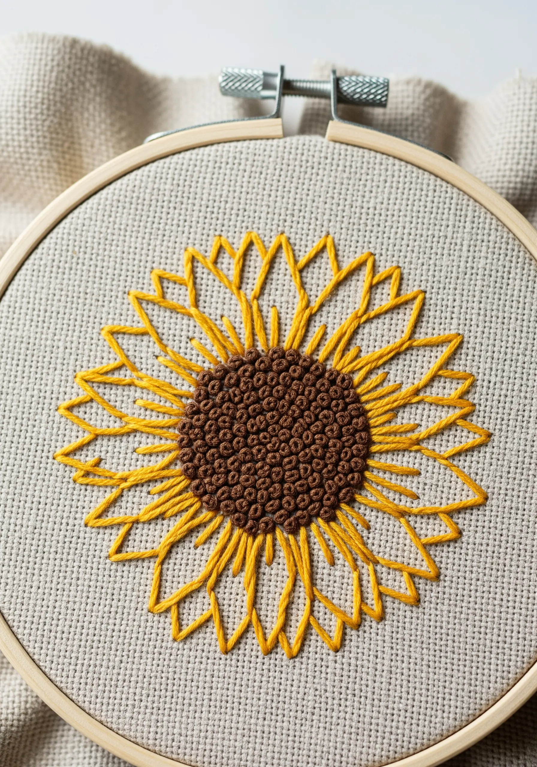 A sunflower with outlined yellow petals and a dense French knot center in a hoop.