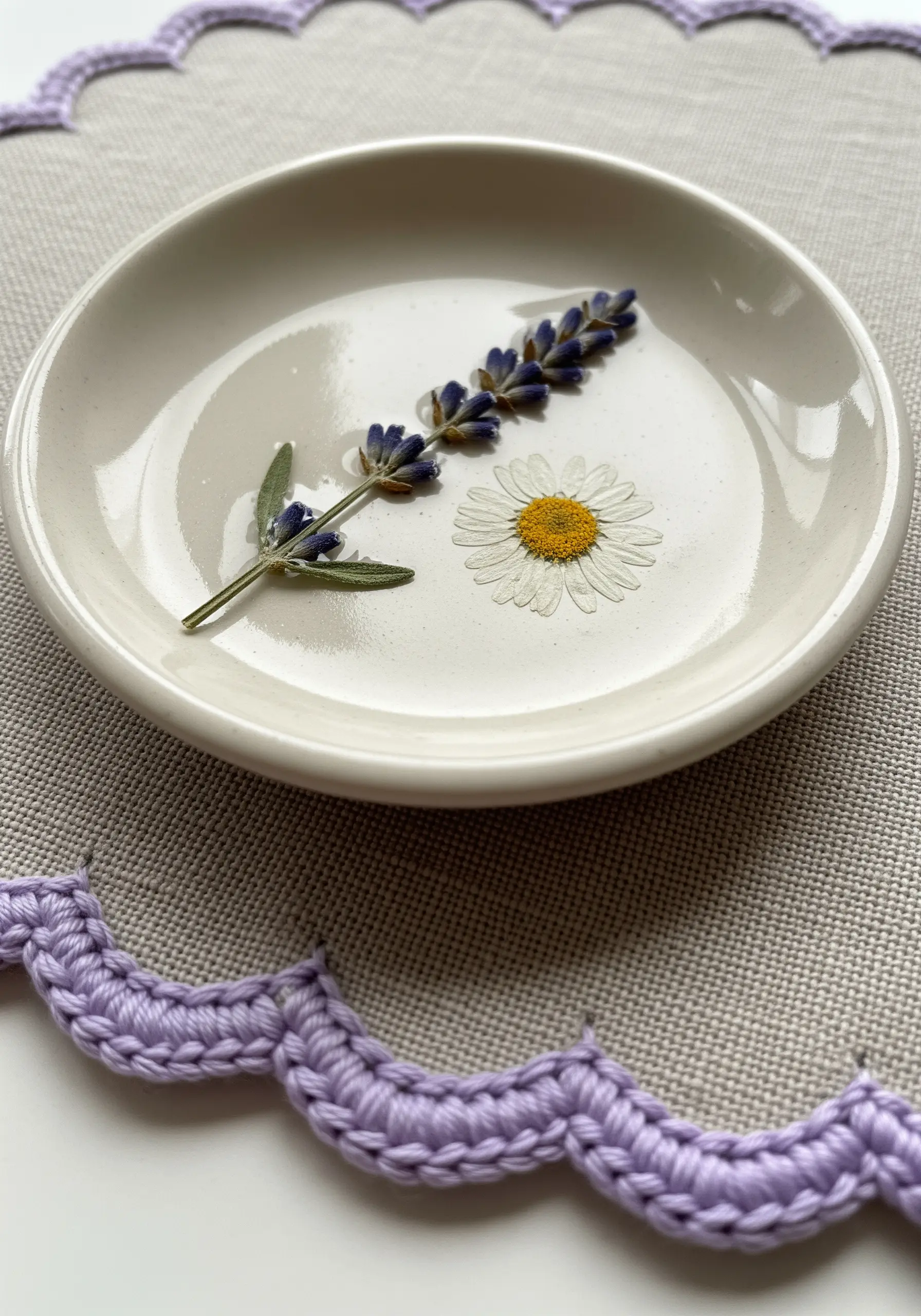 A ceramic dish holding a pressed lavender sprig and a daisy, next to a lavender crocheted border.