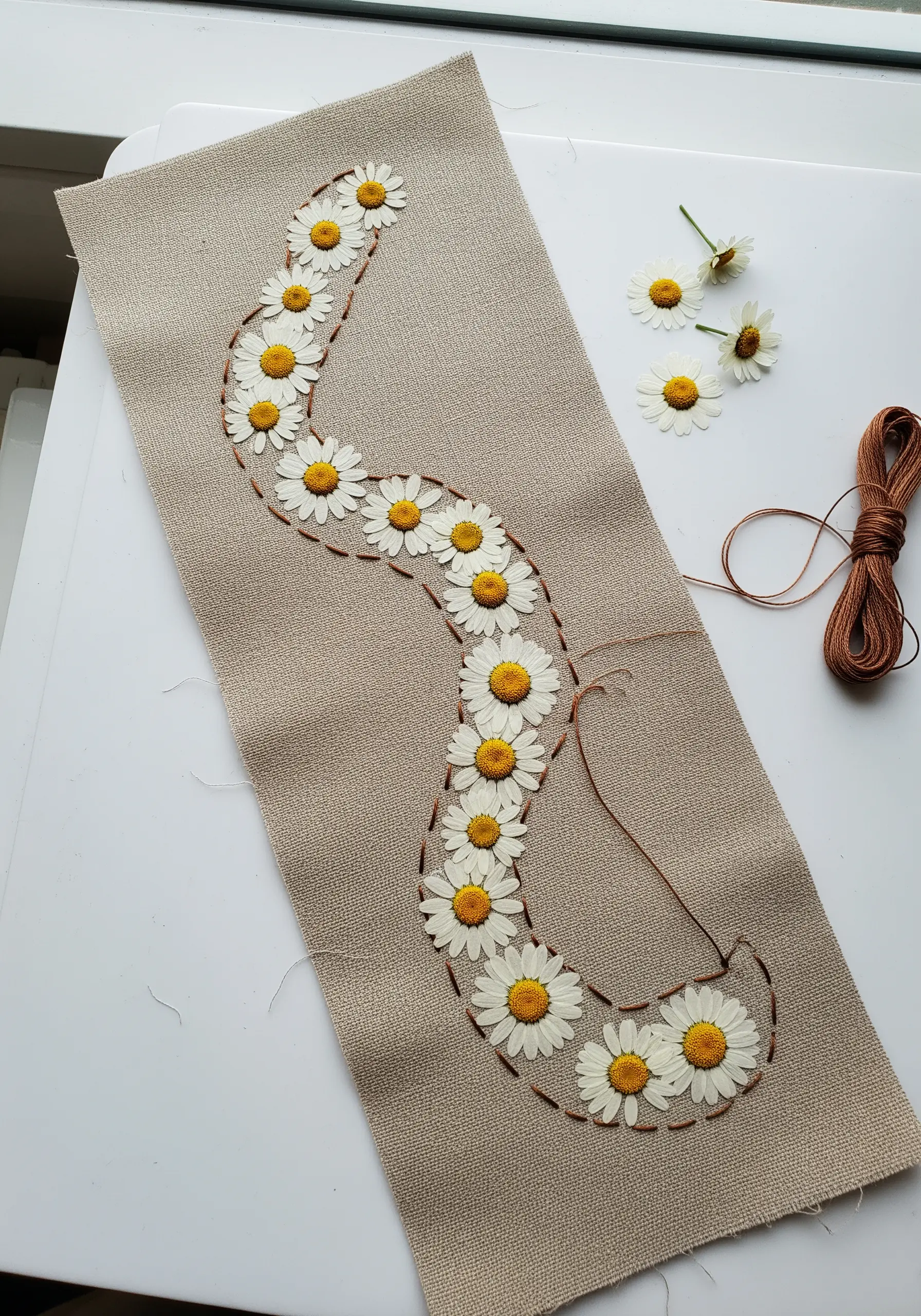 A winding line of pressed daisies stitched onto a neutral linen table runner.