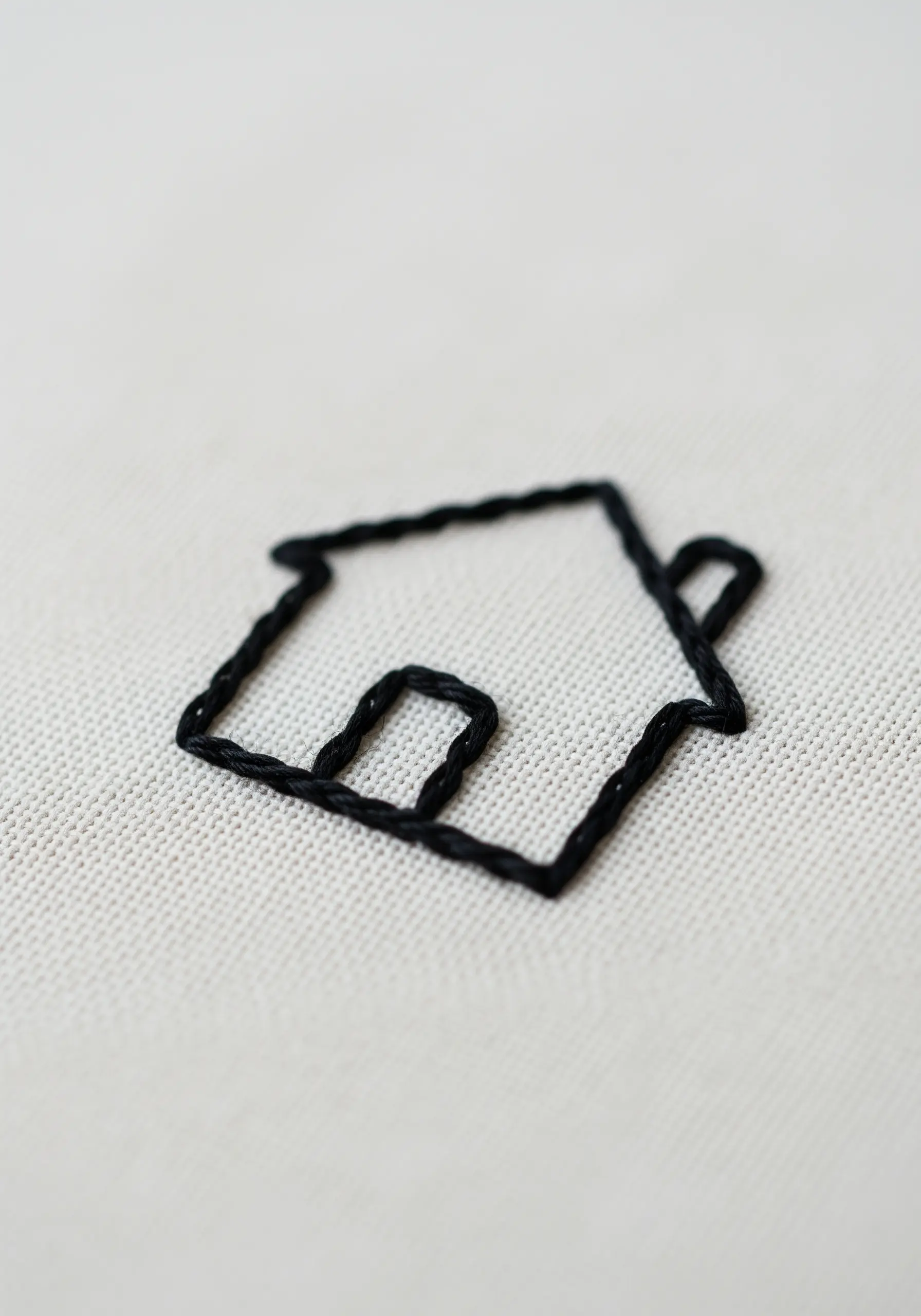 A bold, black outline of a house icon stitched on off-white fabric.