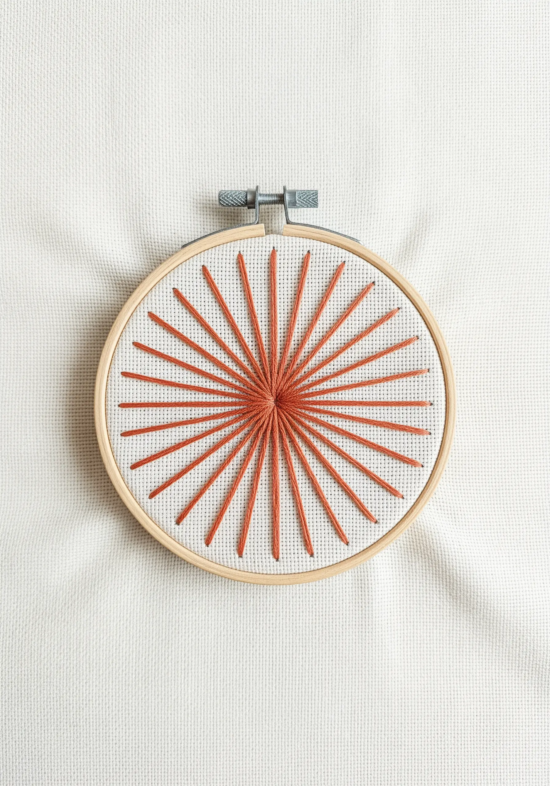 A sunburst design with radiating orange straight stitches on off-white Aida cloth.