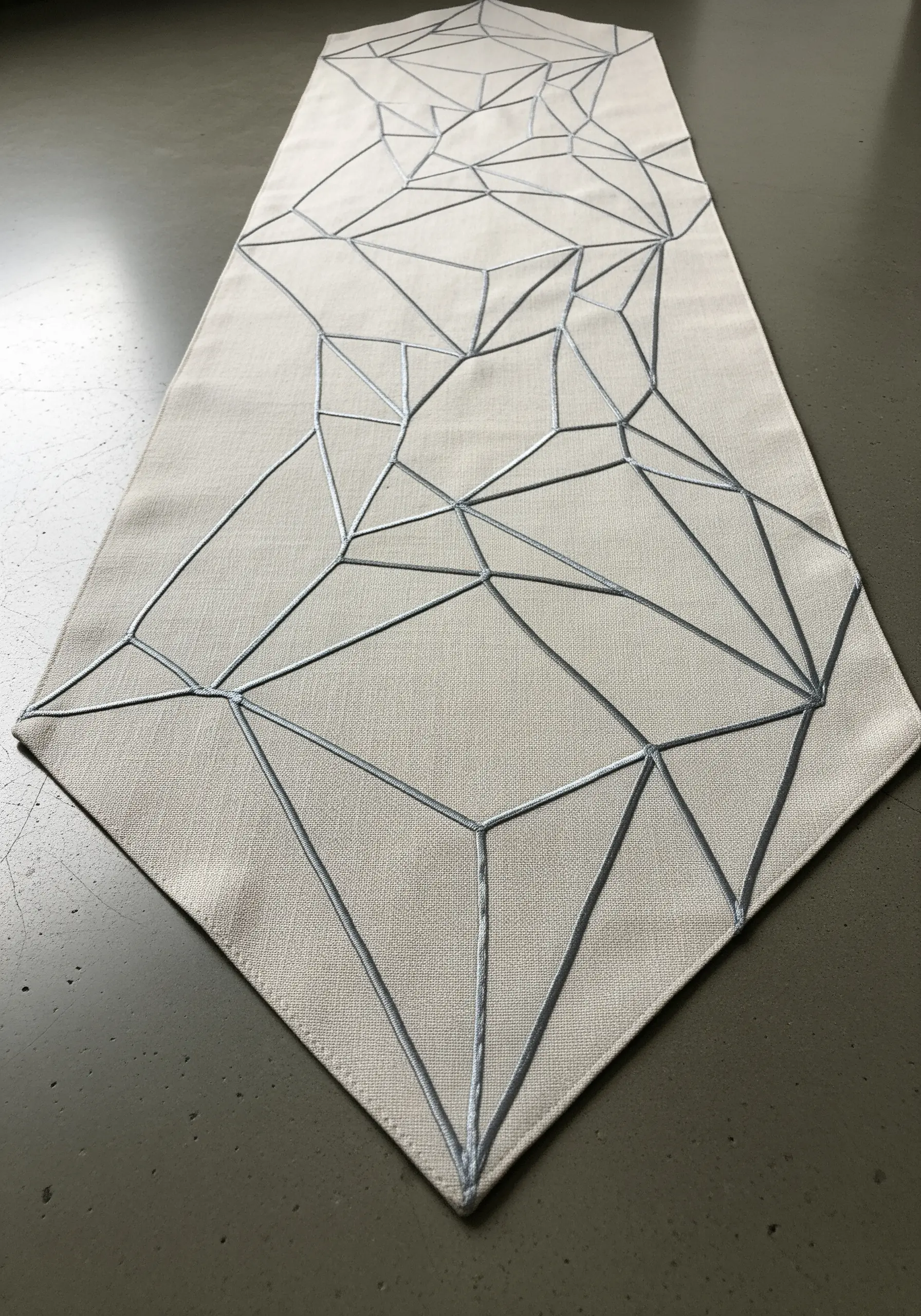 A geometric, web-like pattern of silver lines stitched on a neutral linen table runner.