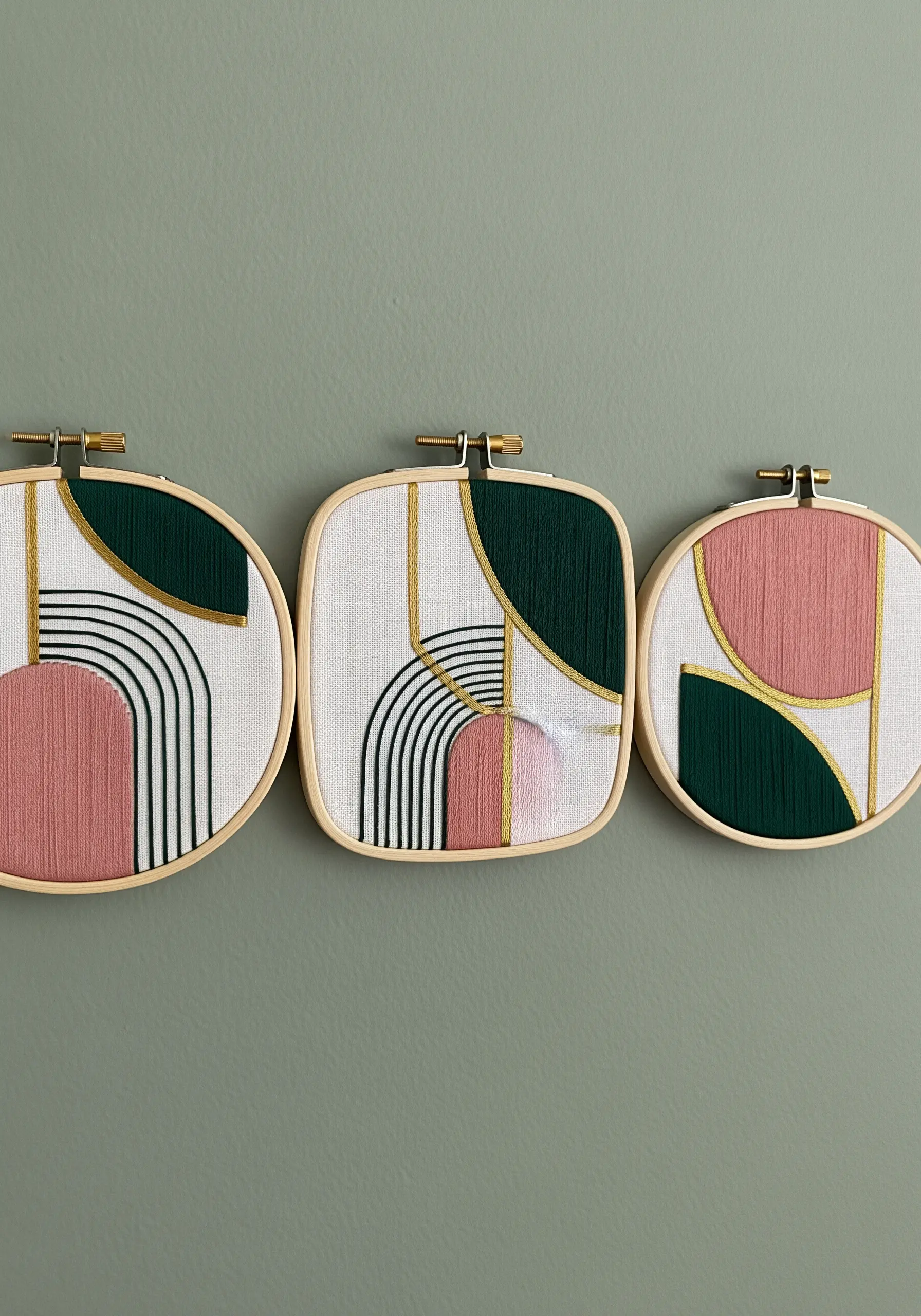 A triptych of embroidery hoops with Art Deco-inspired designs in pink, green, and gold.