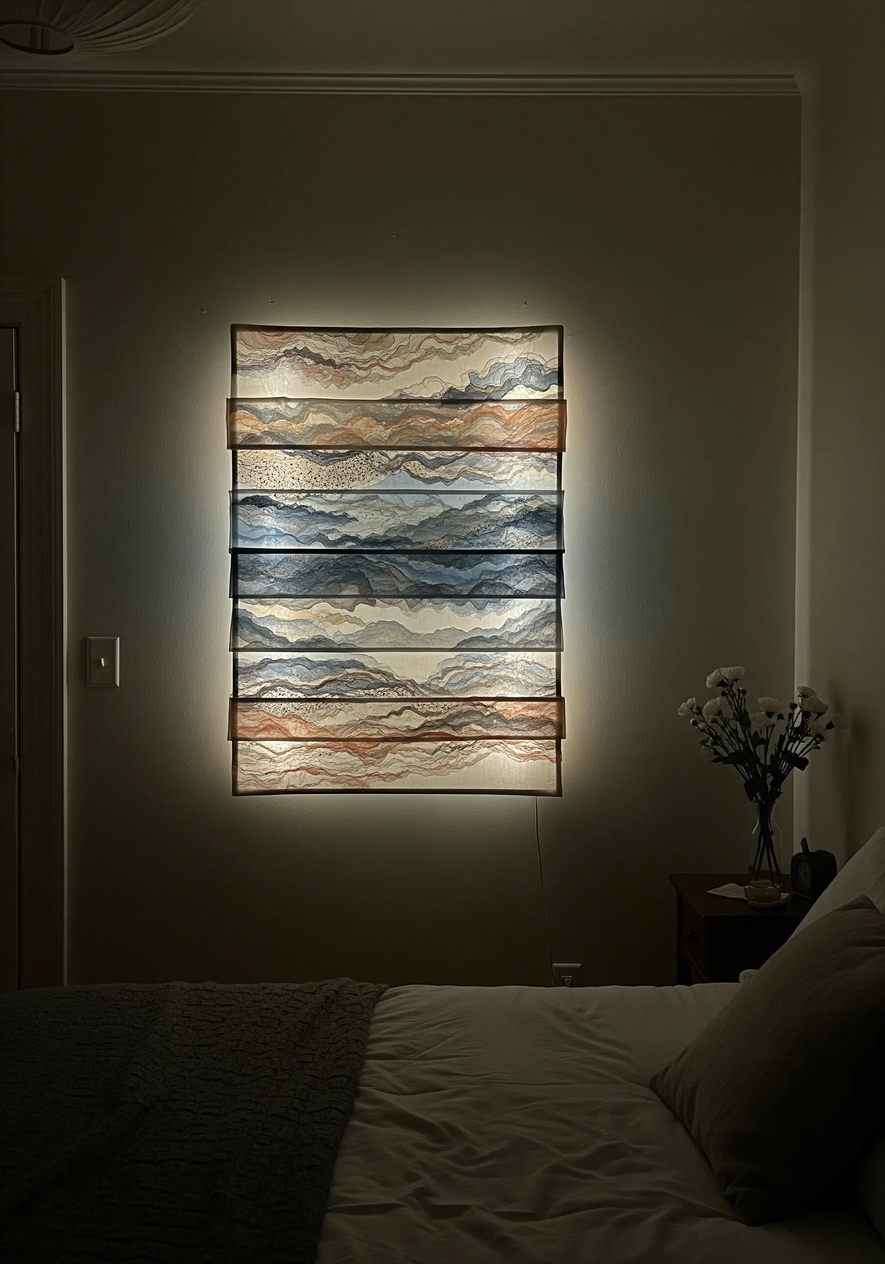A backlit wall hanging made of layered, translucent fabric panels embroidered with an abstract landscape.