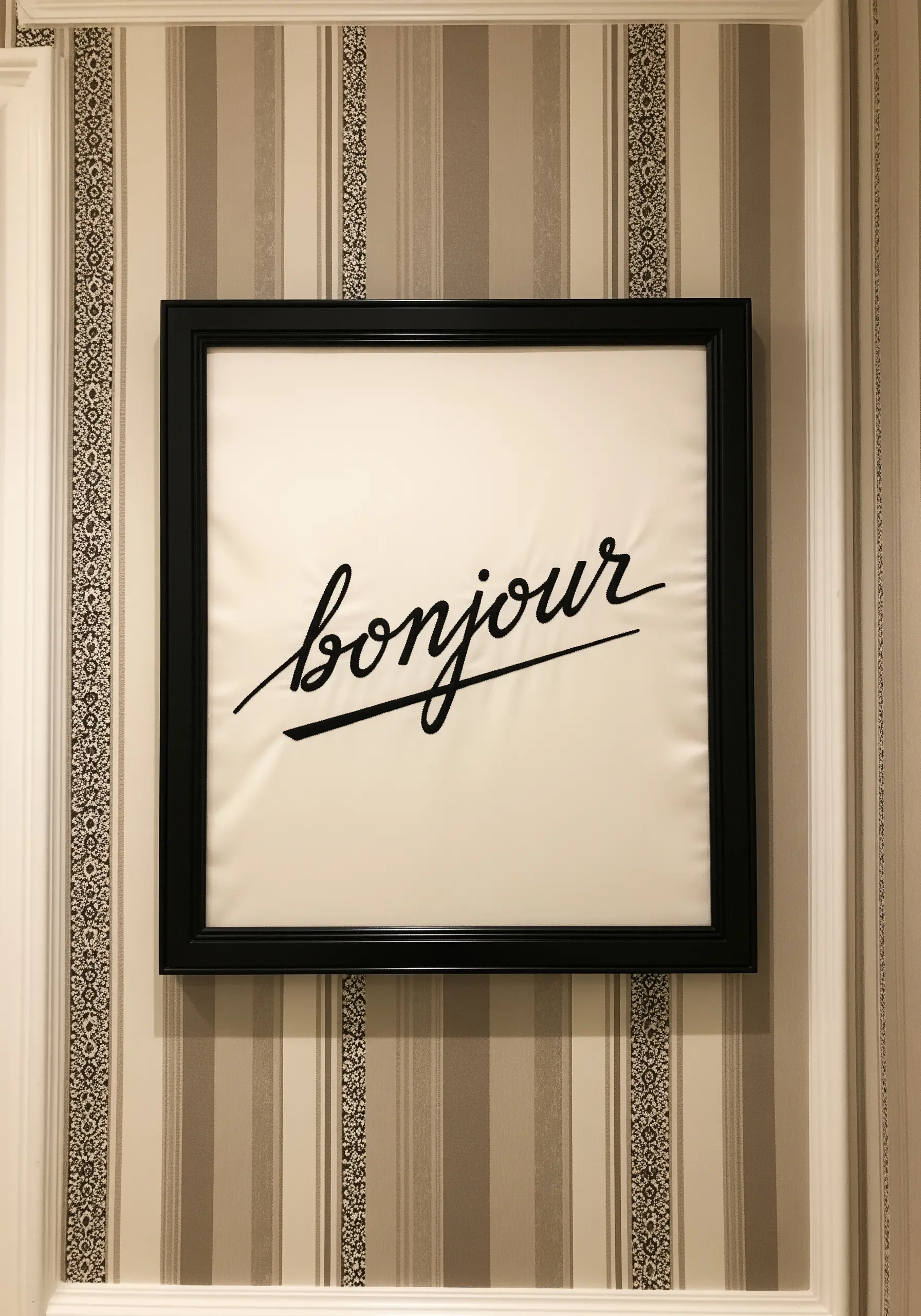 The word 'bonjour' embroidered in a fluid, black script with an underline in a black frame.