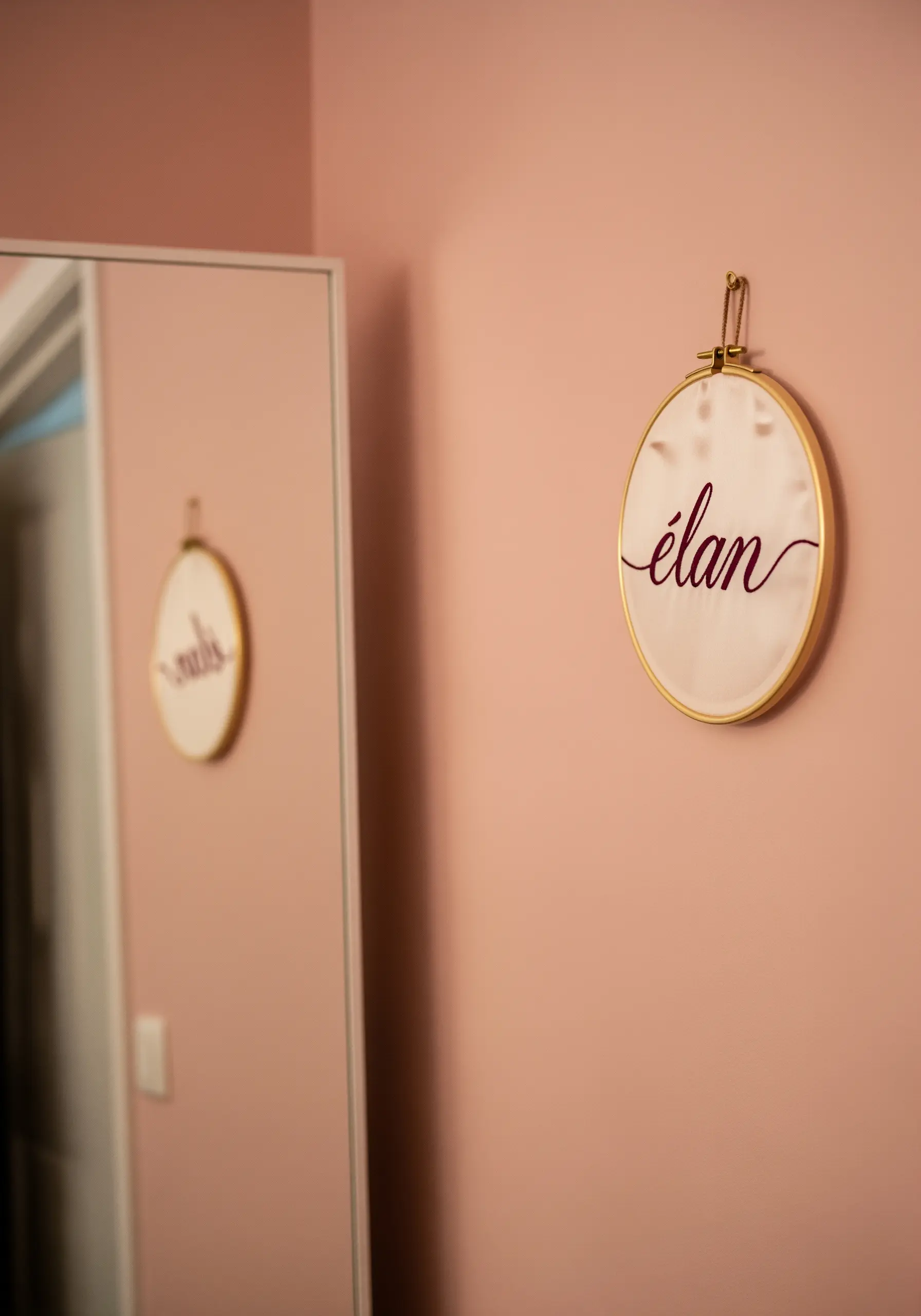 The word 'élan' embroidered in a flowing, burgundy script on light pink fabric in a gold hoop.