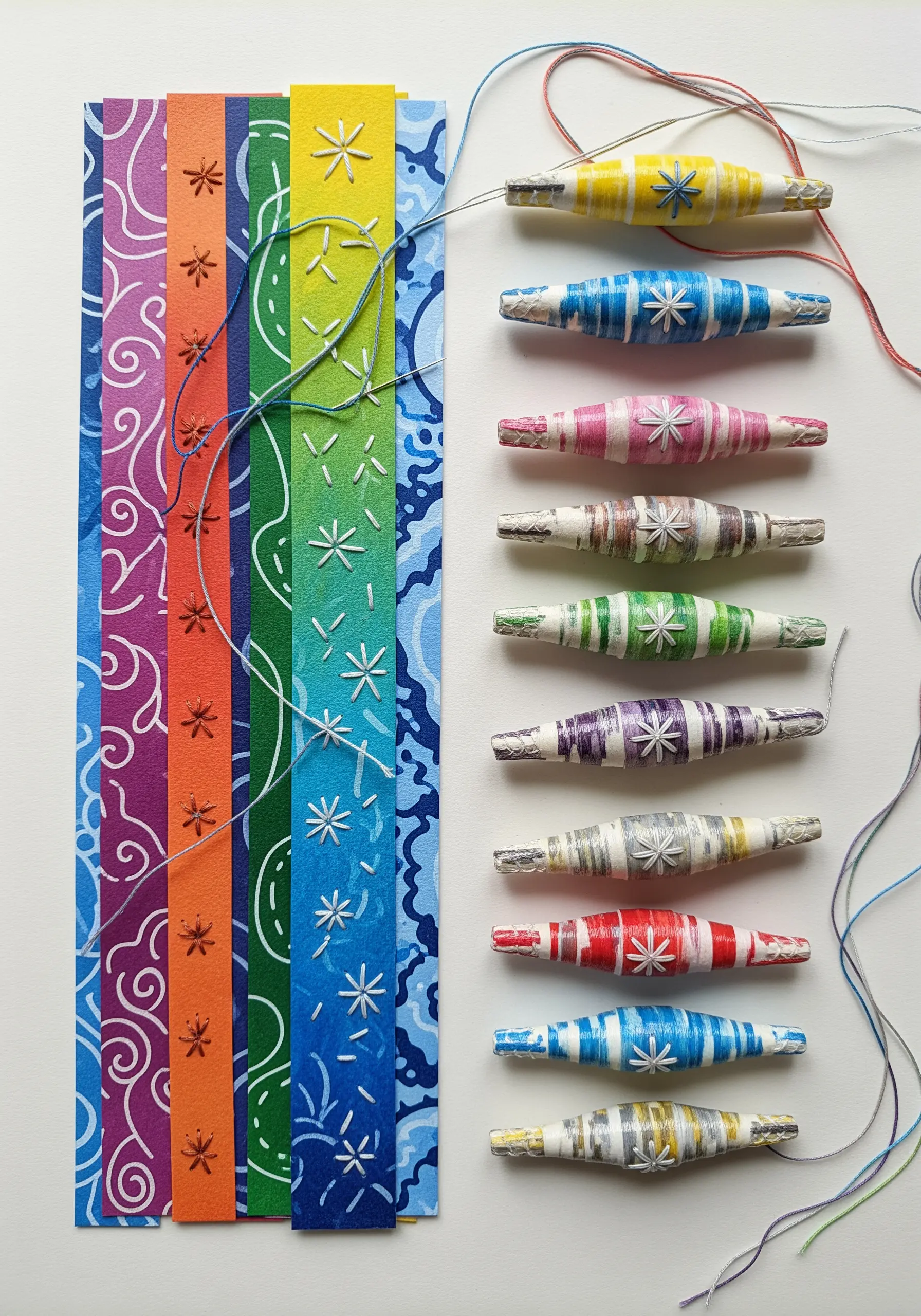 Colorful rolled paper beads decorated with small, embroidered white star stitches.