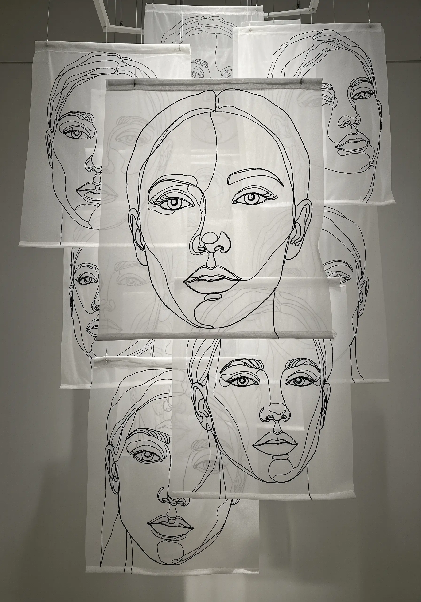 Layered portraits stitched in black thread on sheer white fabric panels, creating a 3D effect.