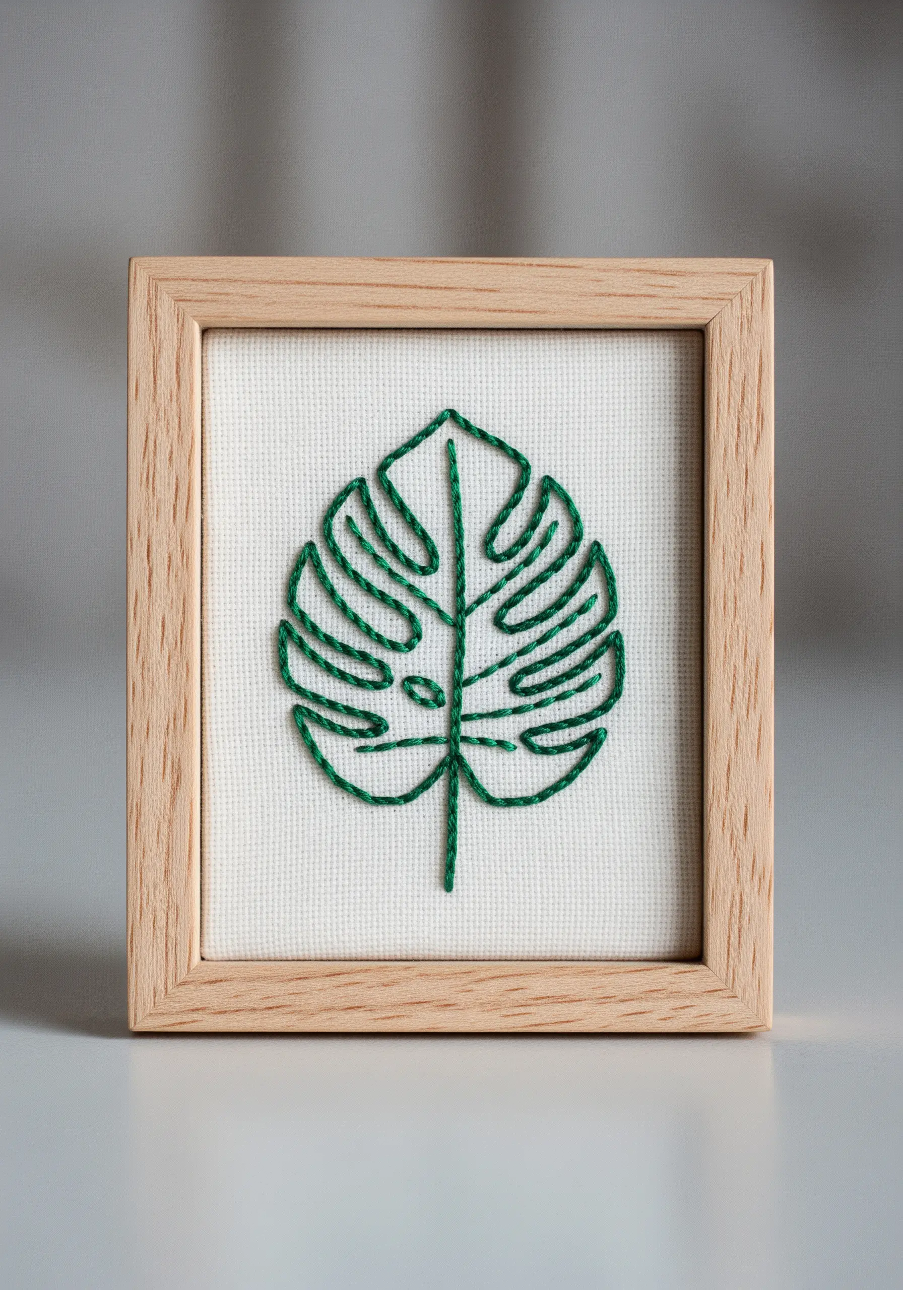 A minimalist outline of a monstera leaf embroidered in green and displayed in a wooden frame.