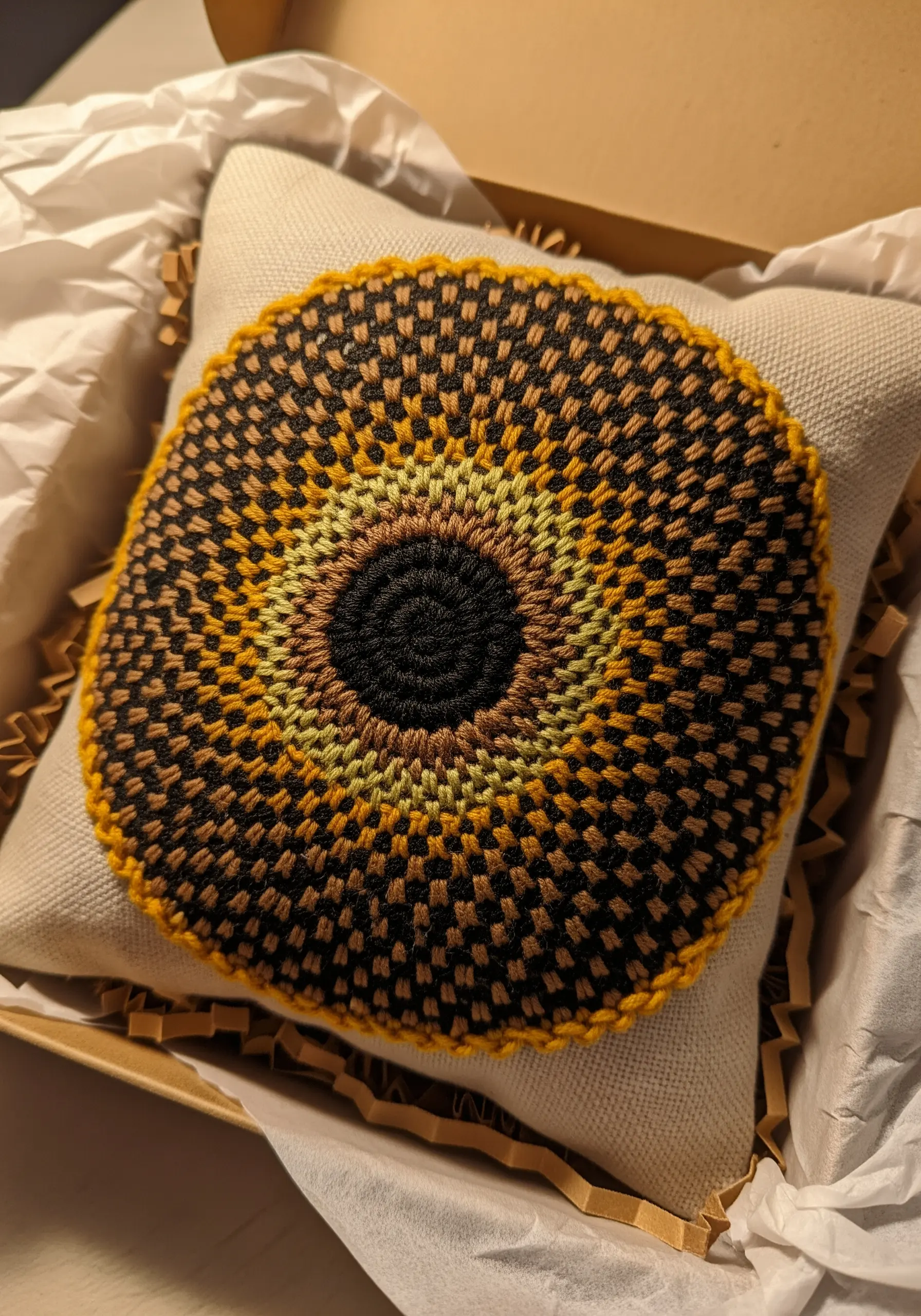 A small decorative pillow embroidered with a geometric checkerboard stitch in brown, yellow, and black.