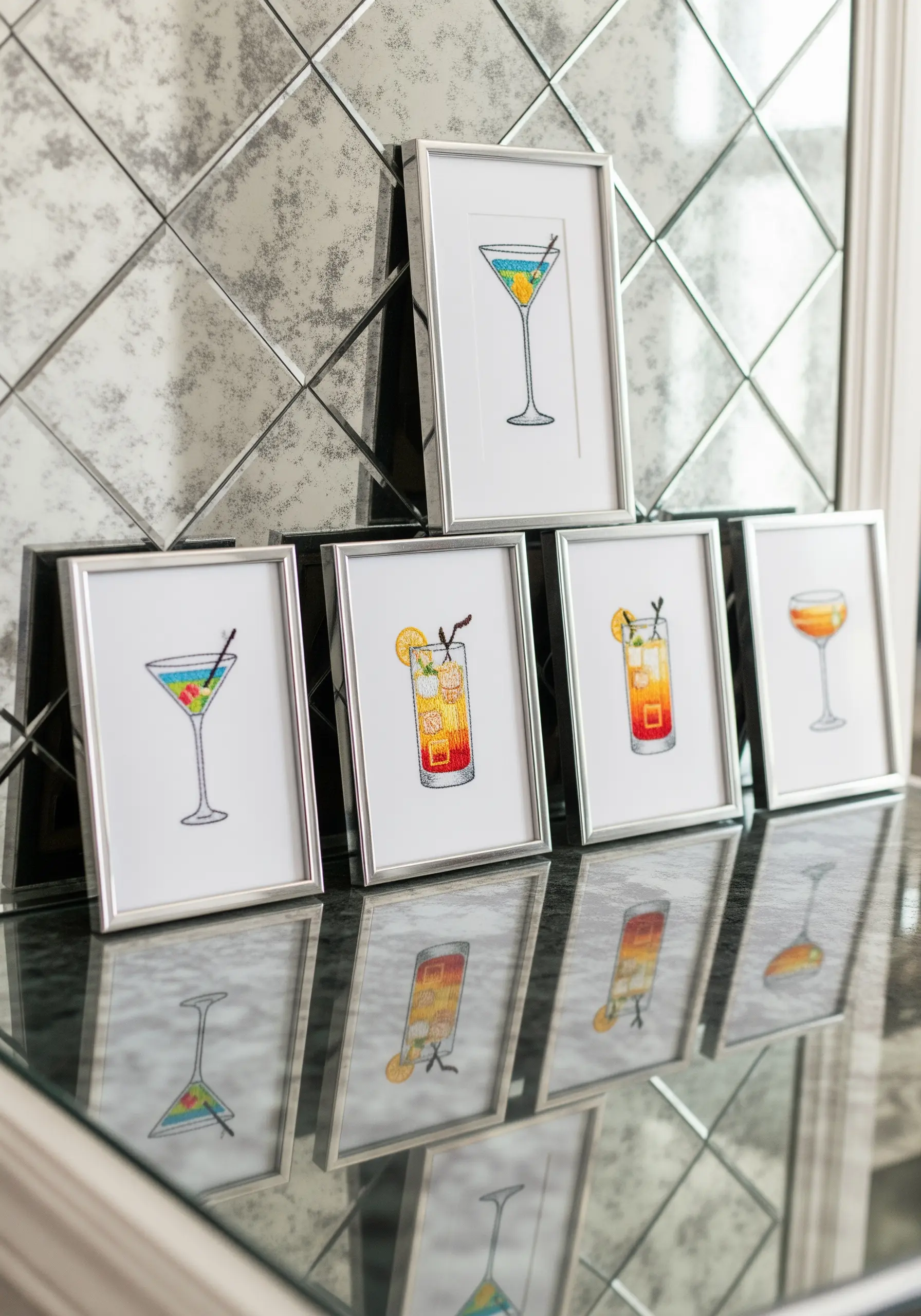 A collection of small framed embroideries of colorful cocktails on a mirrored surface