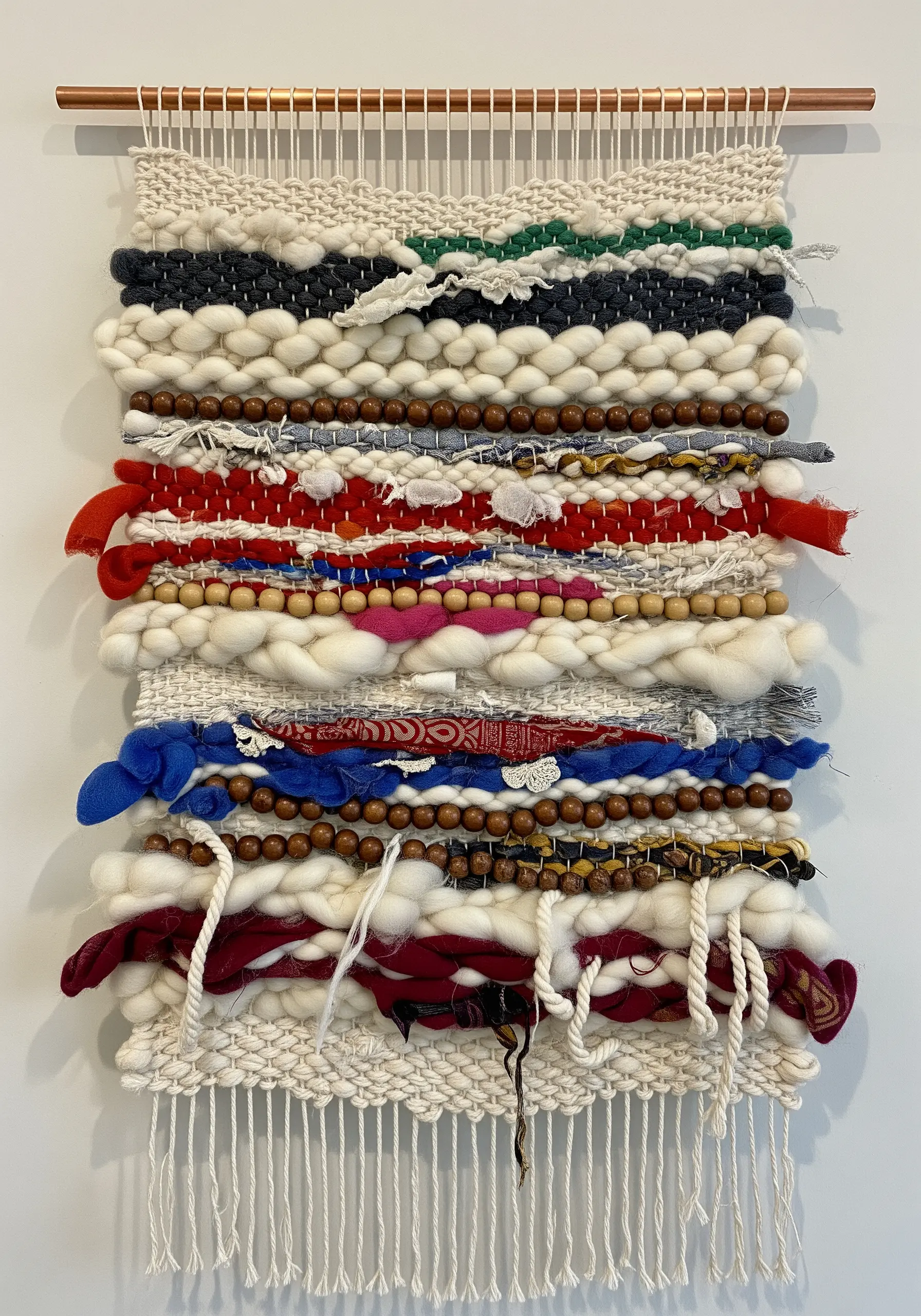 A rustic woven wall hanging incorporating rows of wooden beads and strips of torn fabric.