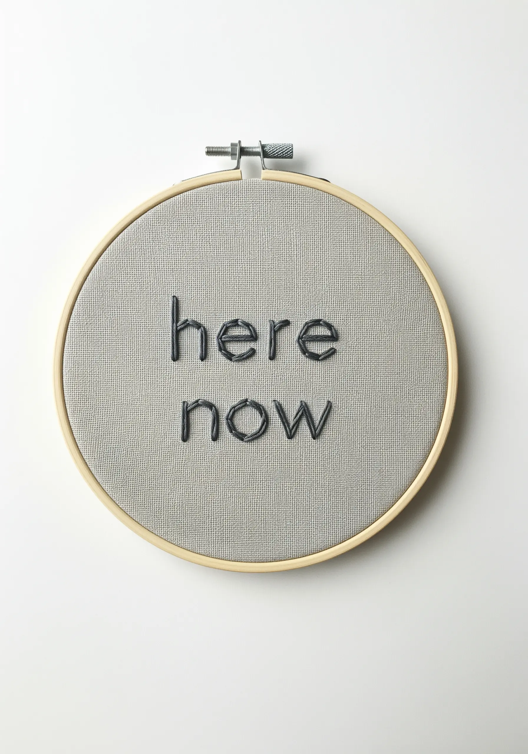 The words 'here now' embroidered in black padded satin stitch on gray linen.