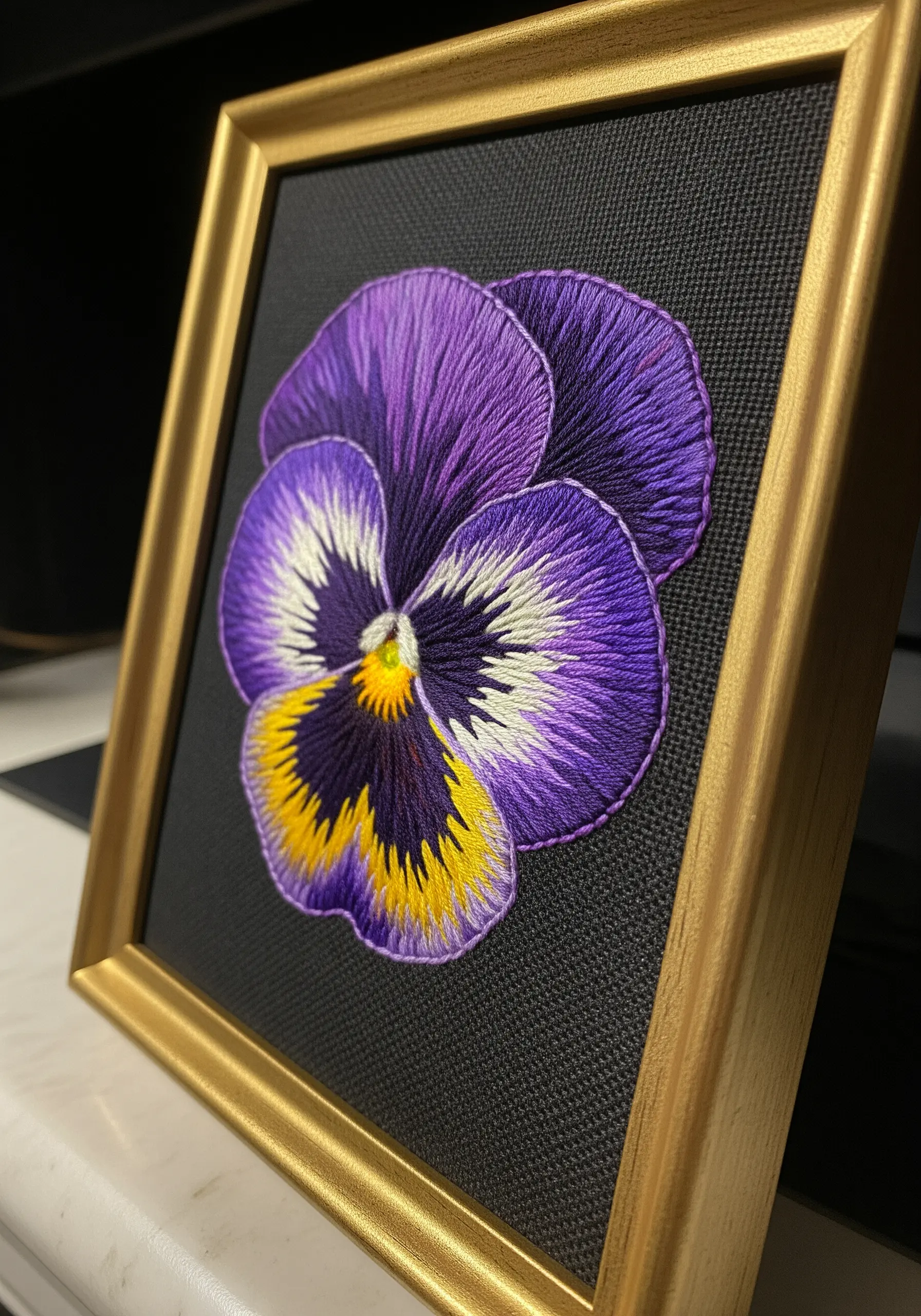 A thread-painted purple, white, and yellow pansy in a gold frame against a black background.