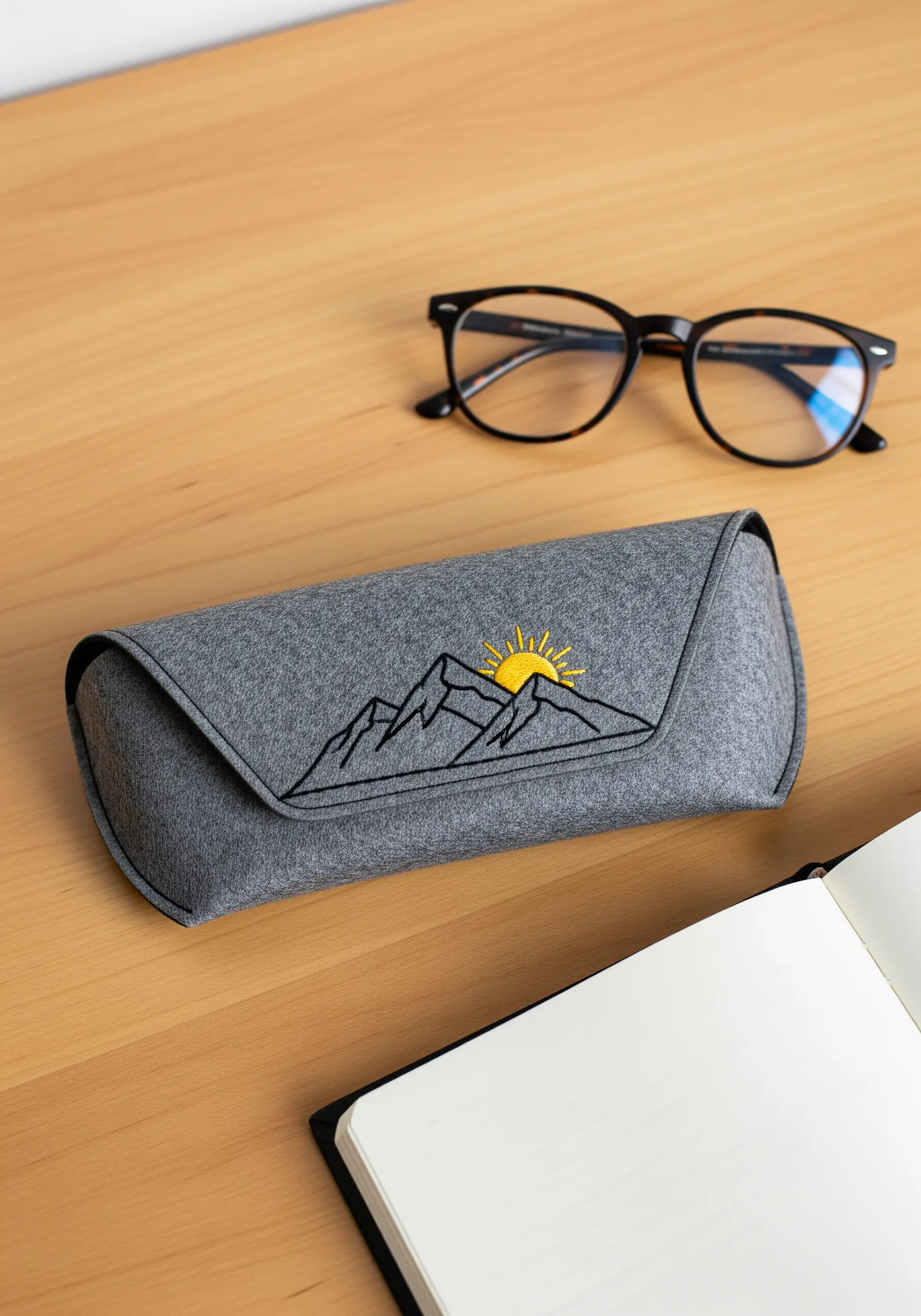A gray felt glasses case embroidered with a minimalist black outline of mountains and a rising sun.