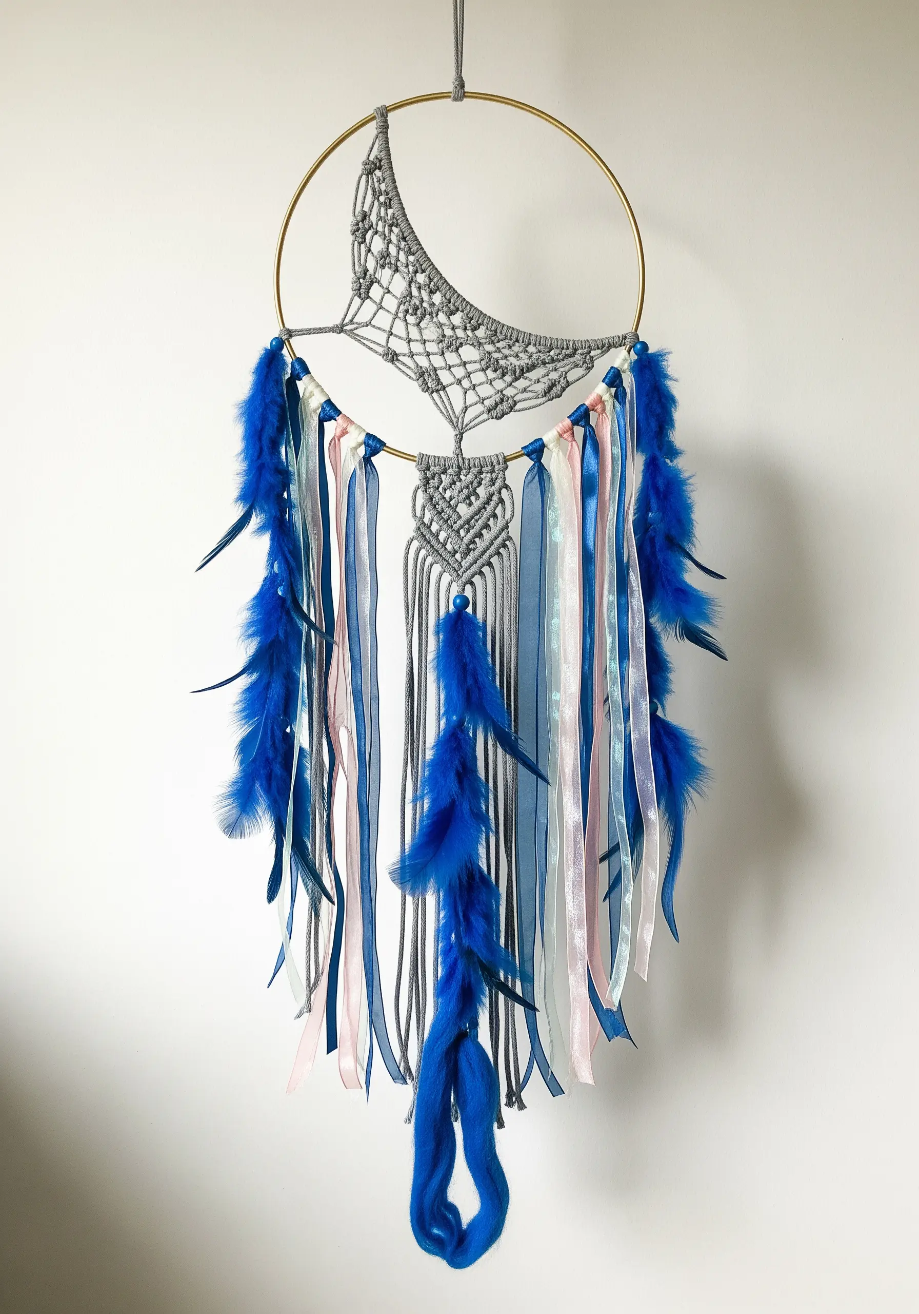 A modern dreamcatcher with a grey macrame moon, blue feathers, and flowing ribbons.