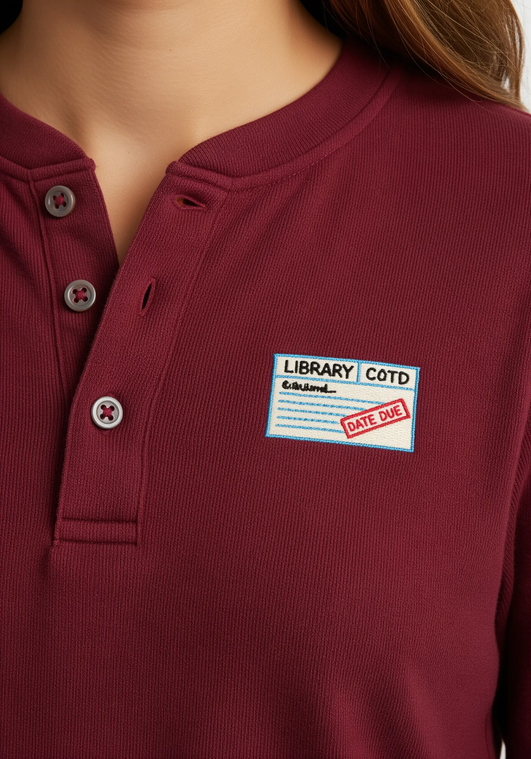 An embroidered patch designed to look like a vintage library card, stitched onto a maroon shirt.