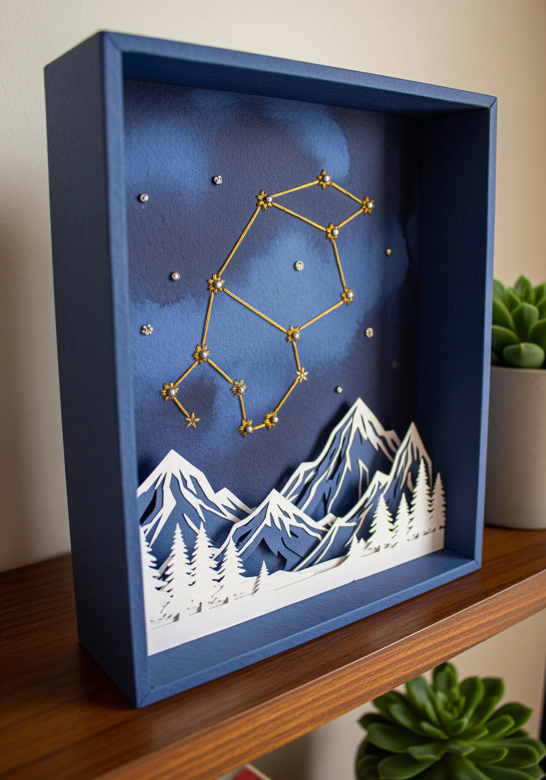 A shadow box with layered paper mountains and a gold-thread constellation against a night sky.