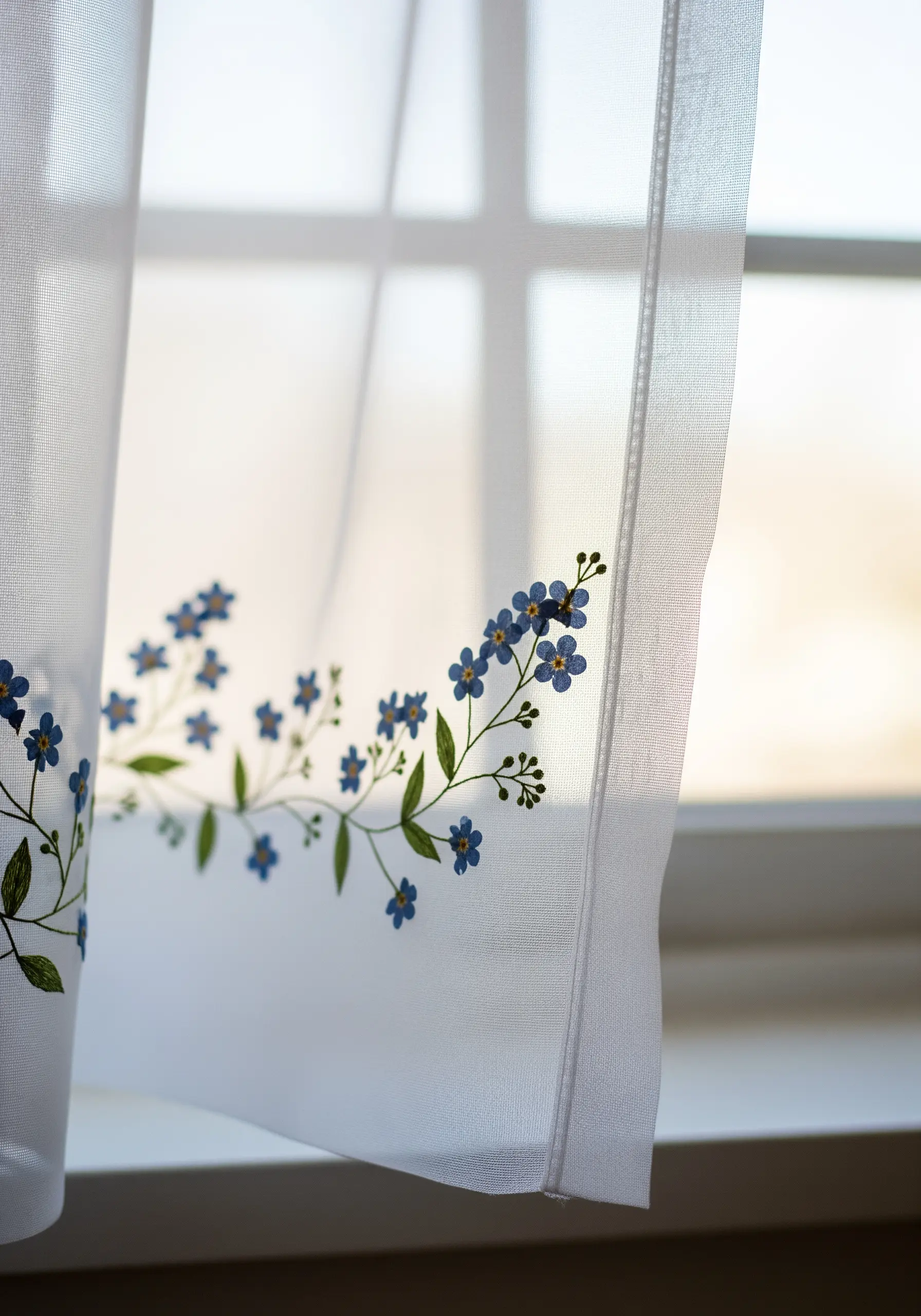 Delicate blue forget-me-not flowers embroidered on the hem of a sheer white curtain.