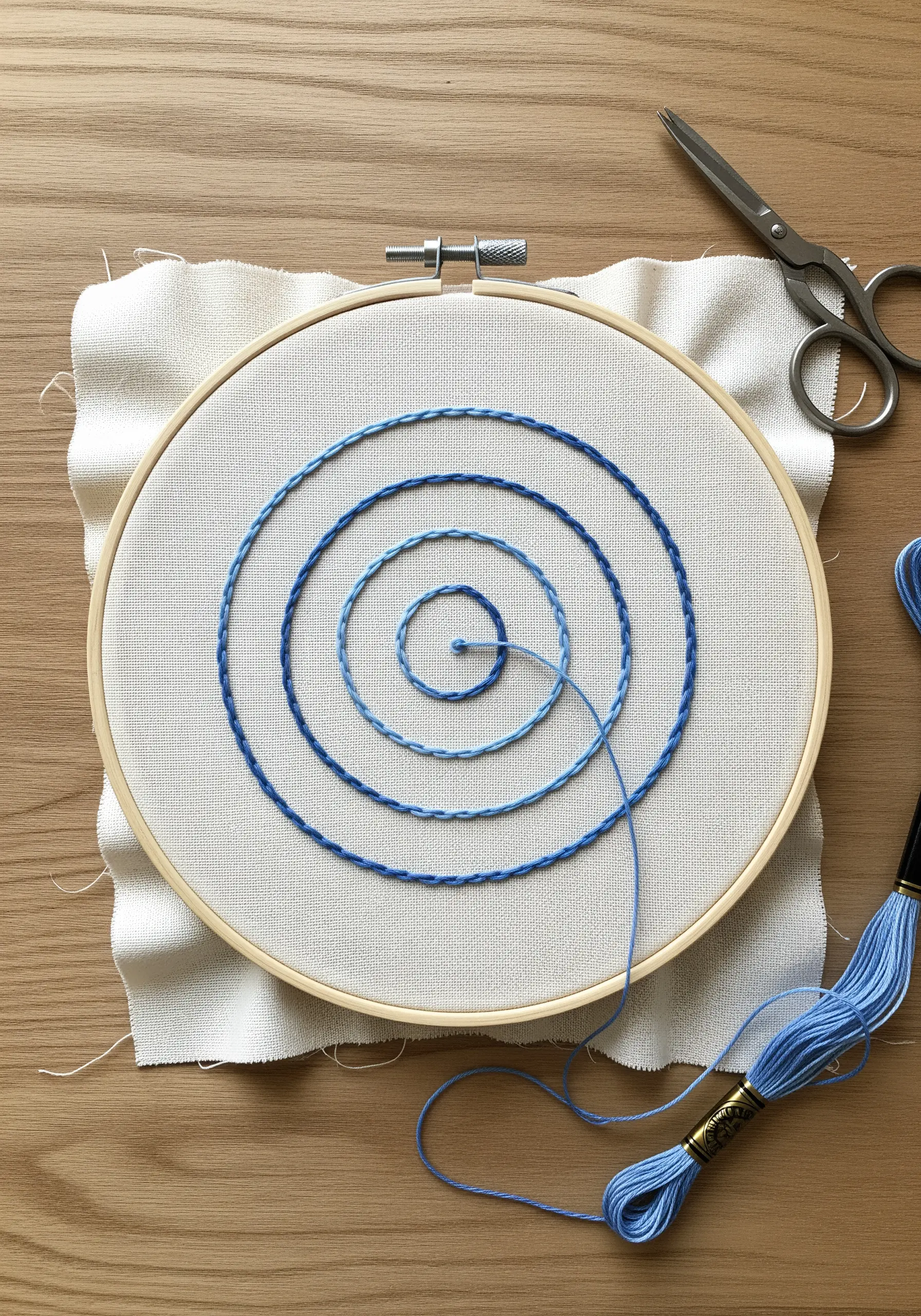 An embroidery hoop showing a spiral stitched with a gradient of blue threads on white fabric.
