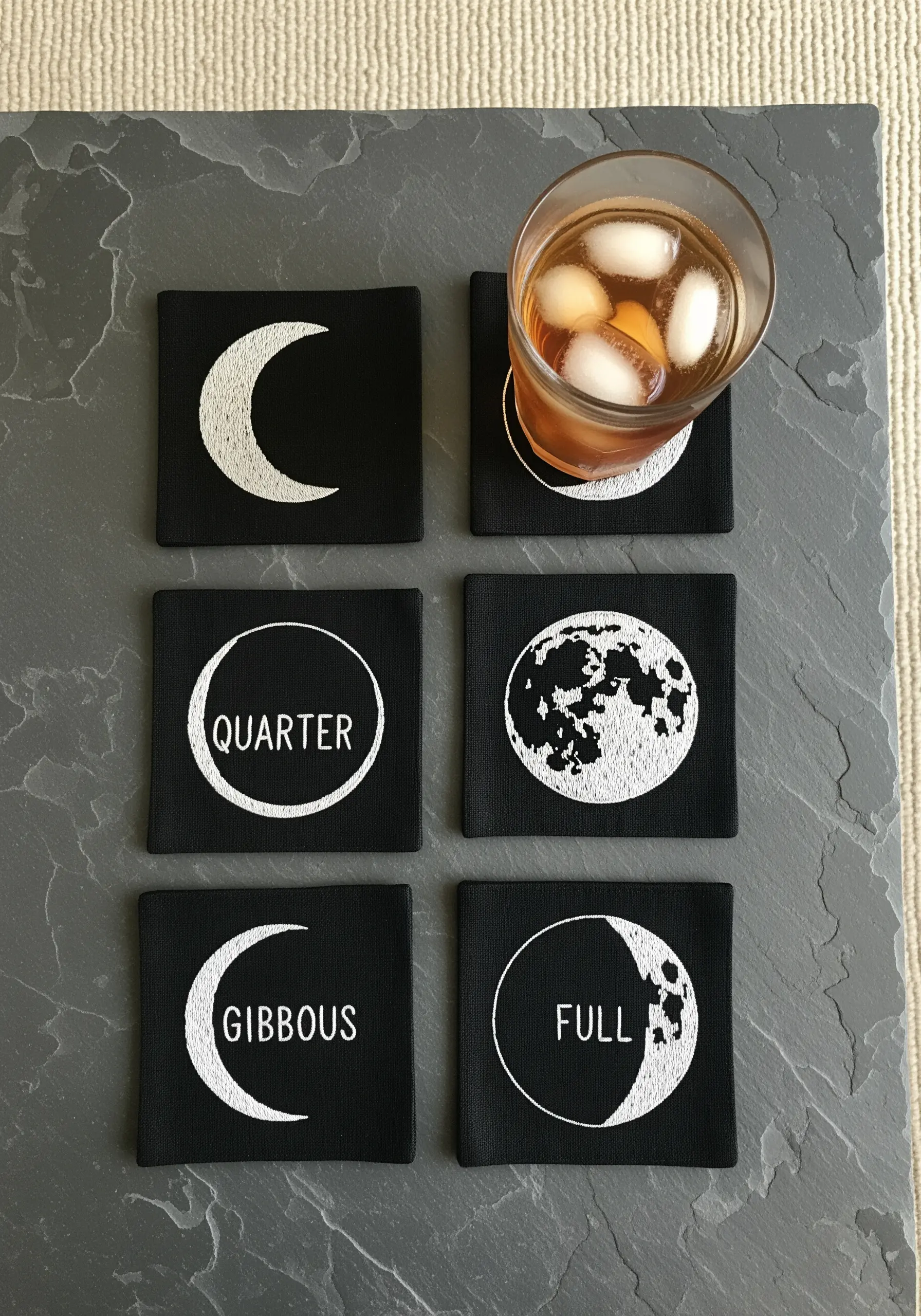 A set of six black coasters, each embroidered with a different phase of the moon in white.