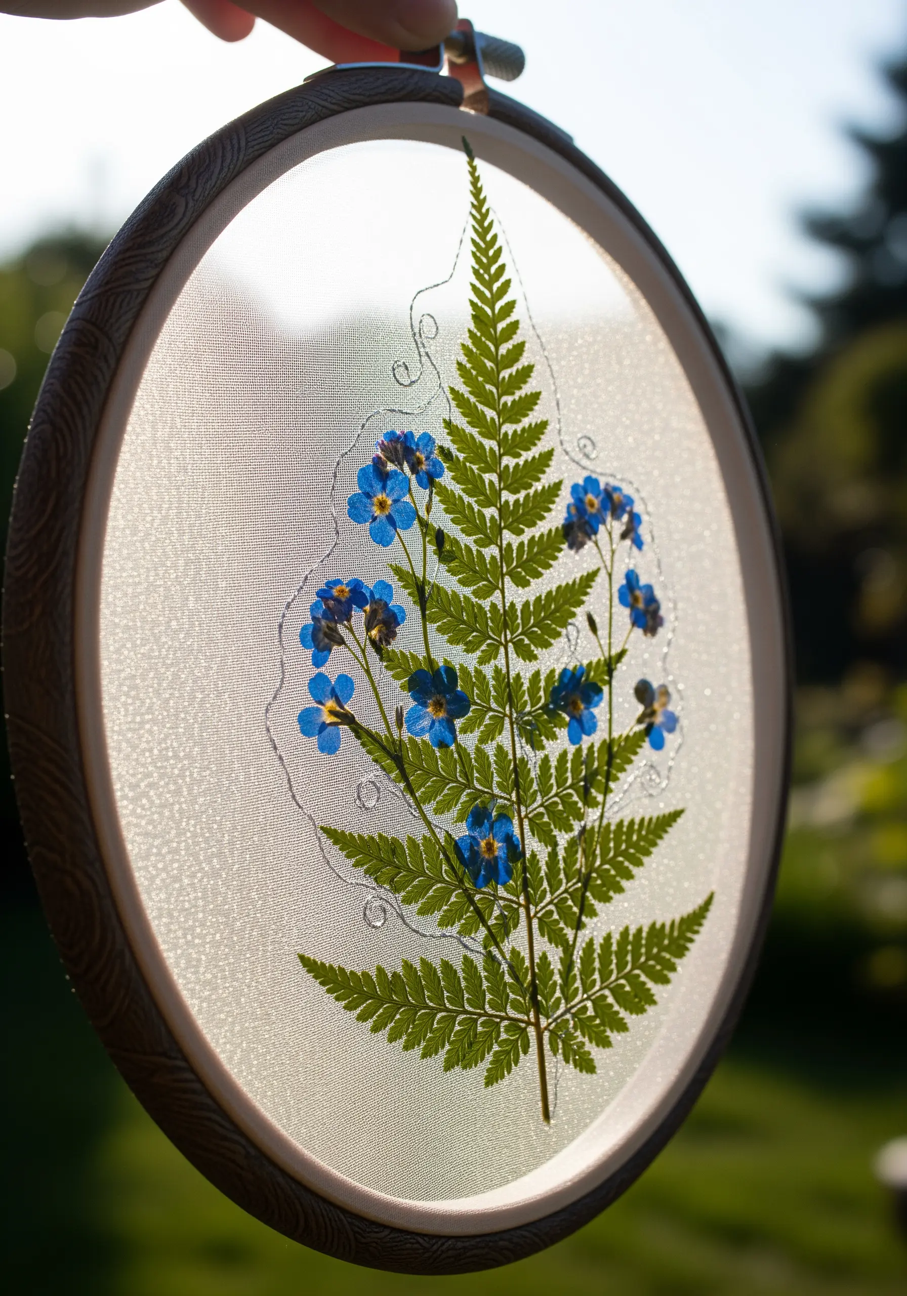 A pressed fern and blue forget-me-nots stitched onto sheer fabric in an embroidery hoop.