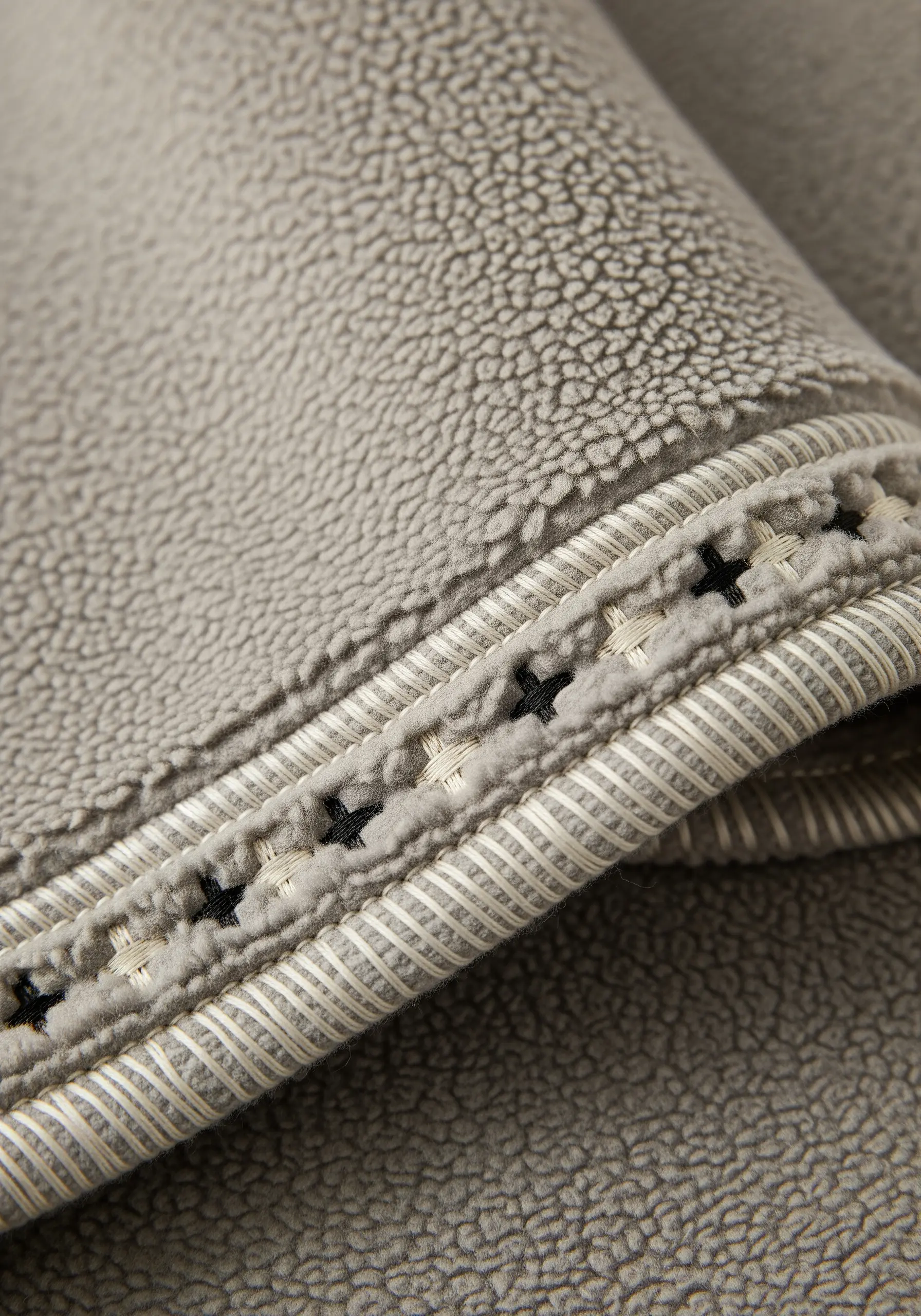 Black cross-stitch and cream running stitch detail along the hem of a grey fleece blanket.