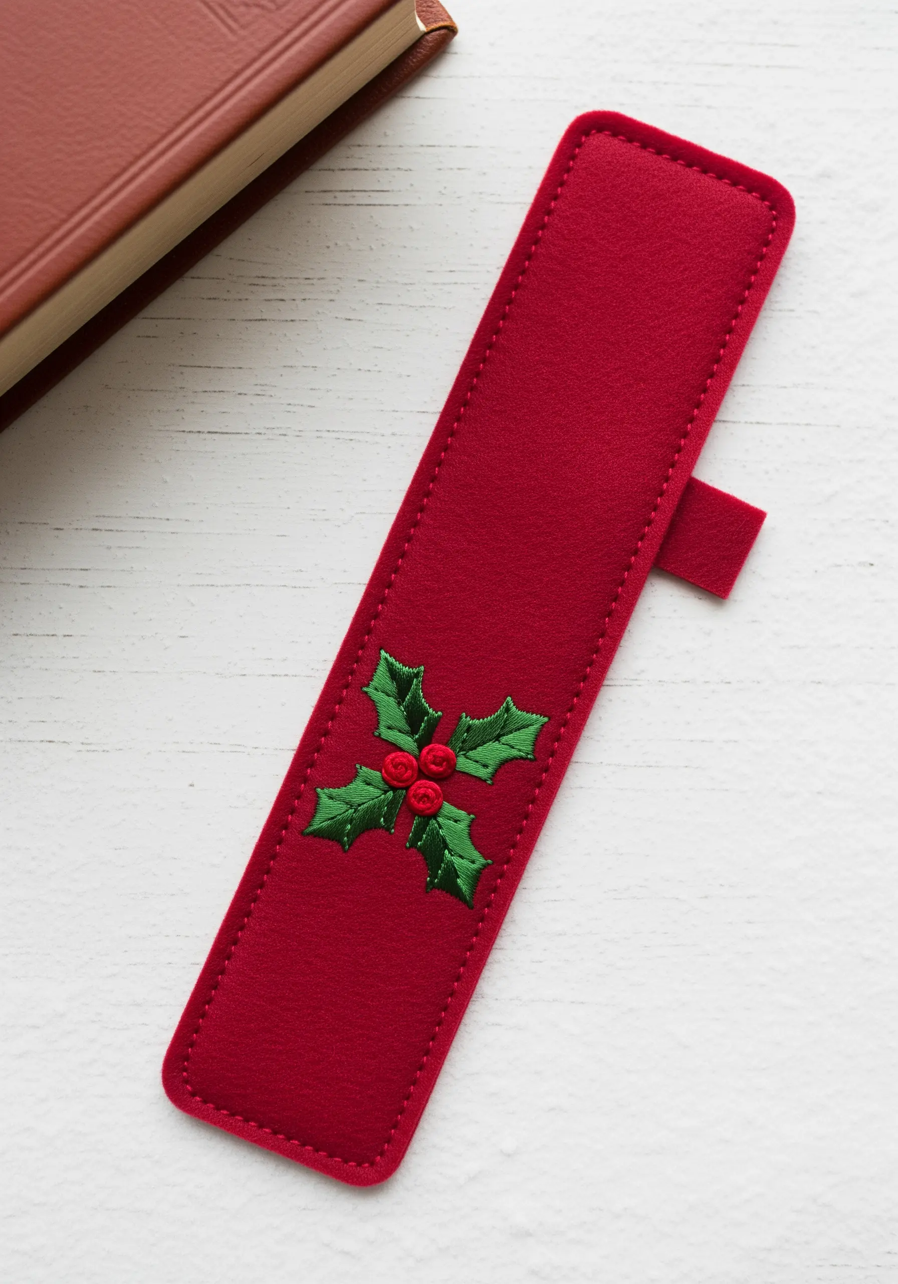 A red felt bookmark sleeve decorated with a small embroidered holly and berry motif.