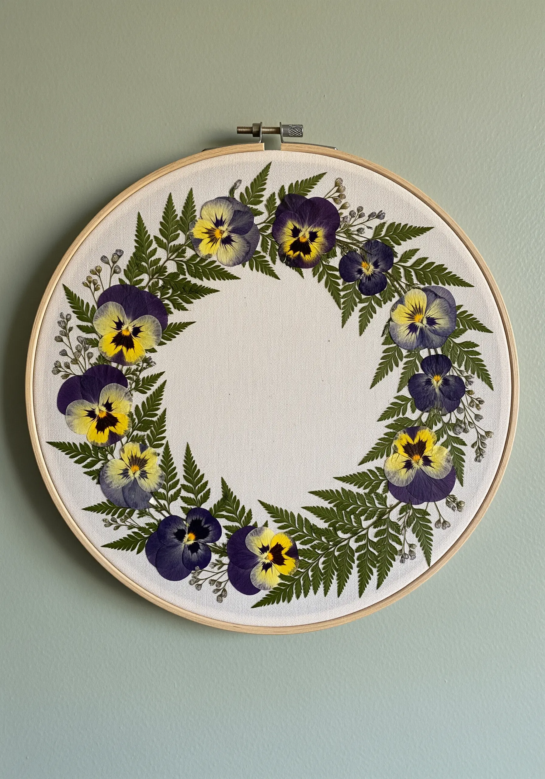 An embroidery hoop with a wreath of real pressed pansies and ferns on off-white fabric.
