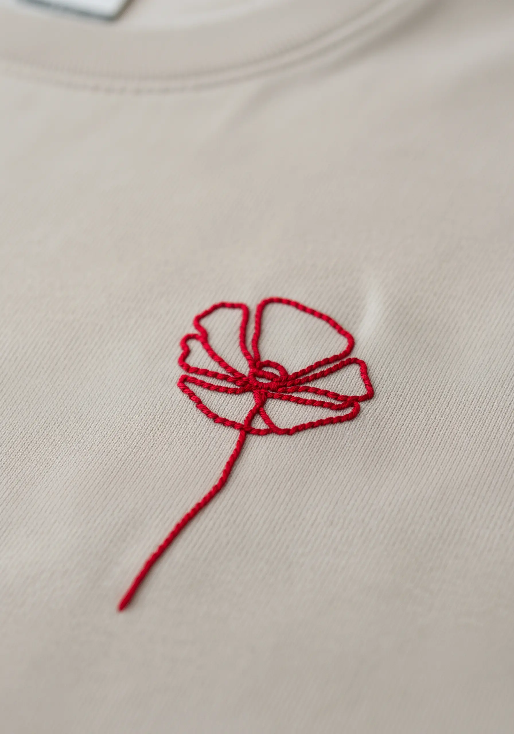 A single red thread creates a minimalist outline of a poppy on a beige t-shirt.