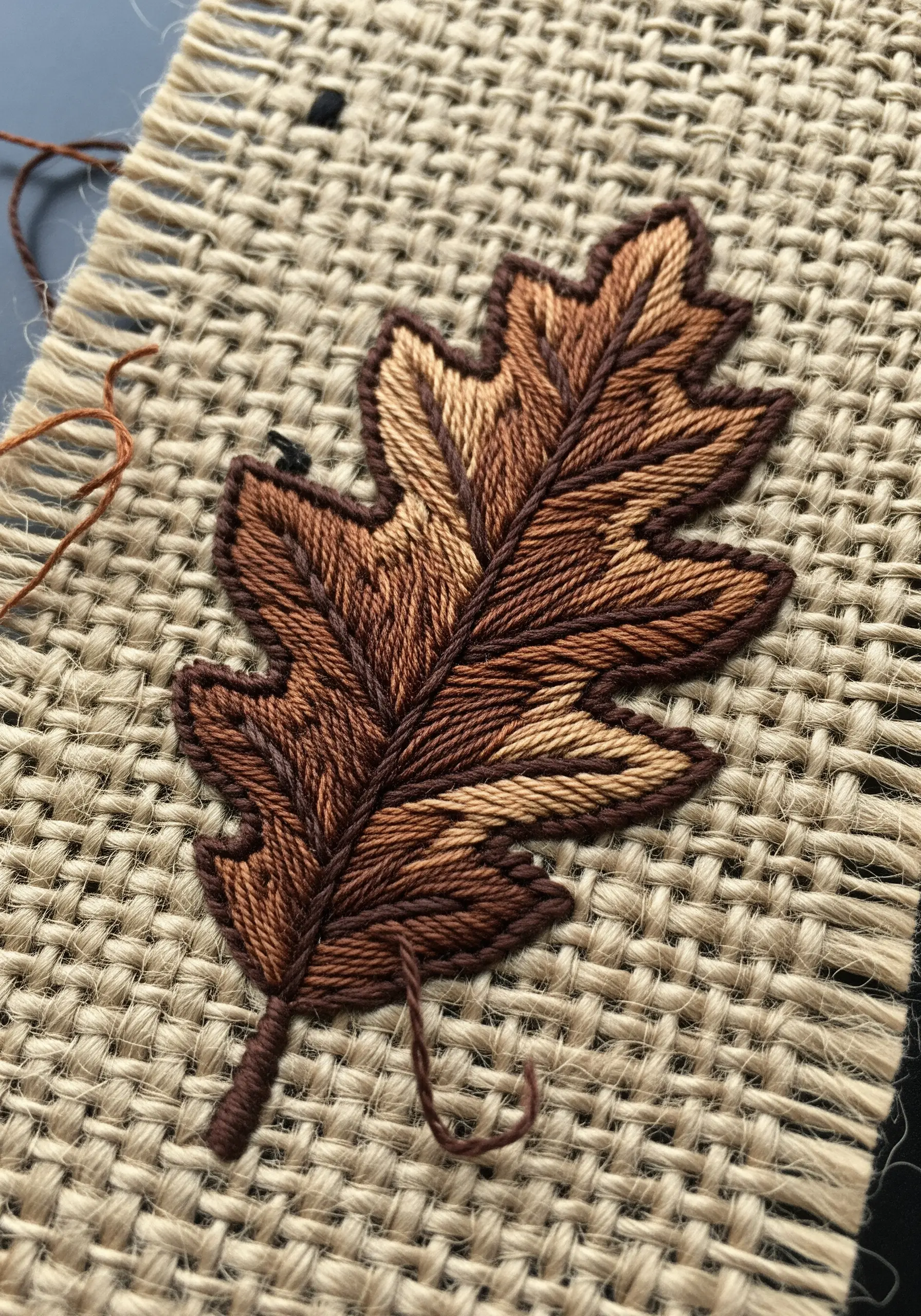 A detailed oak leaf embroidered on coarse burlap fabric with blended brown threads.