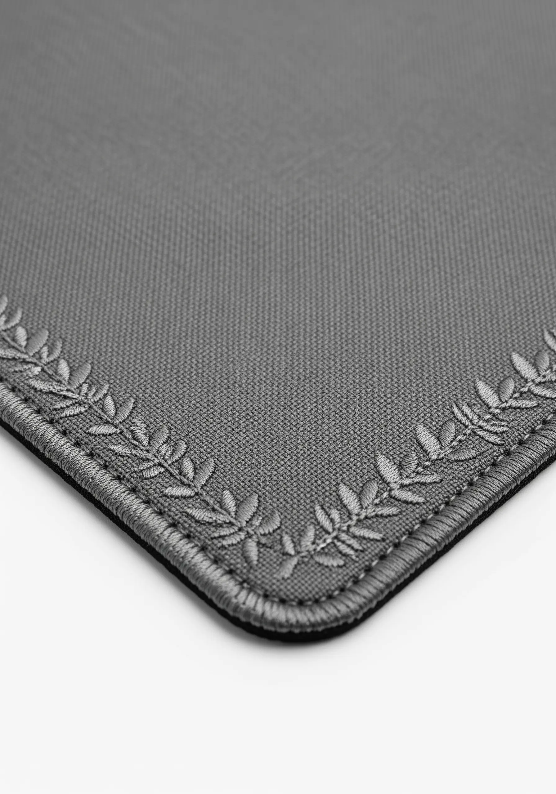 A grey desk mat with a subtle, tone-on-tone embroidered laurel leaf border in the corner.