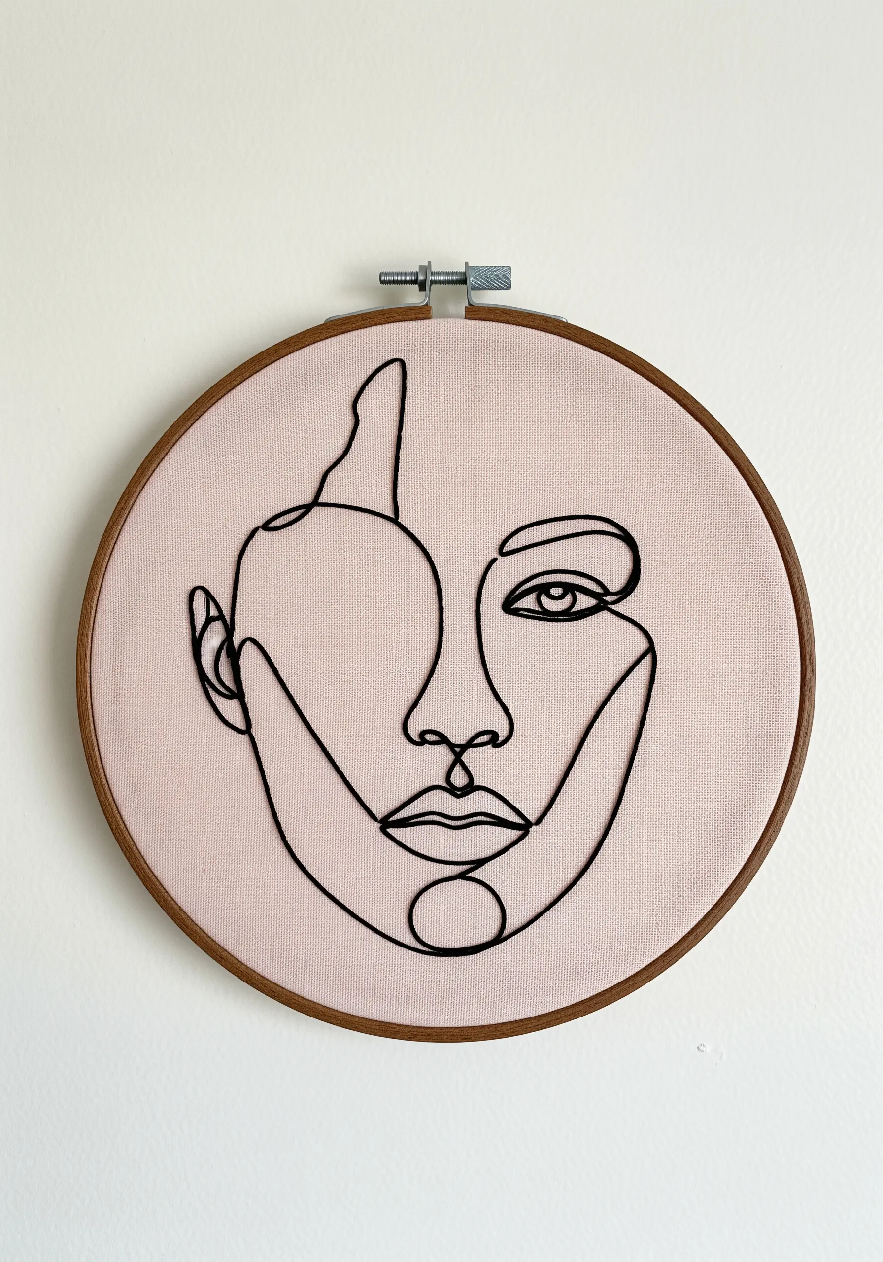 Minimalist single-line face portrait embroidered in black thread on pink fabric.