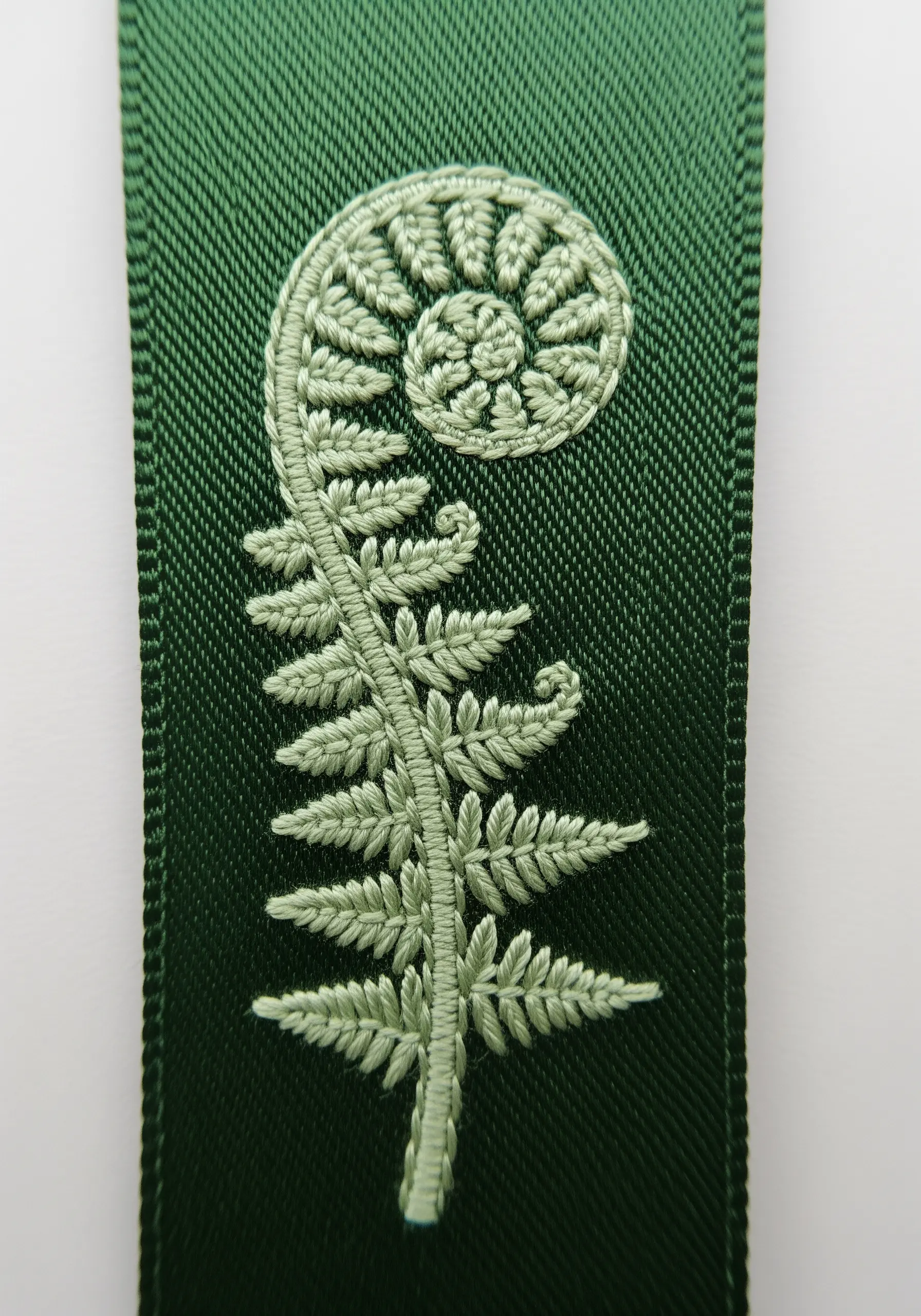 A detailed, unfurling fiddlehead fern embroidered in light green on a dark green ribbon.