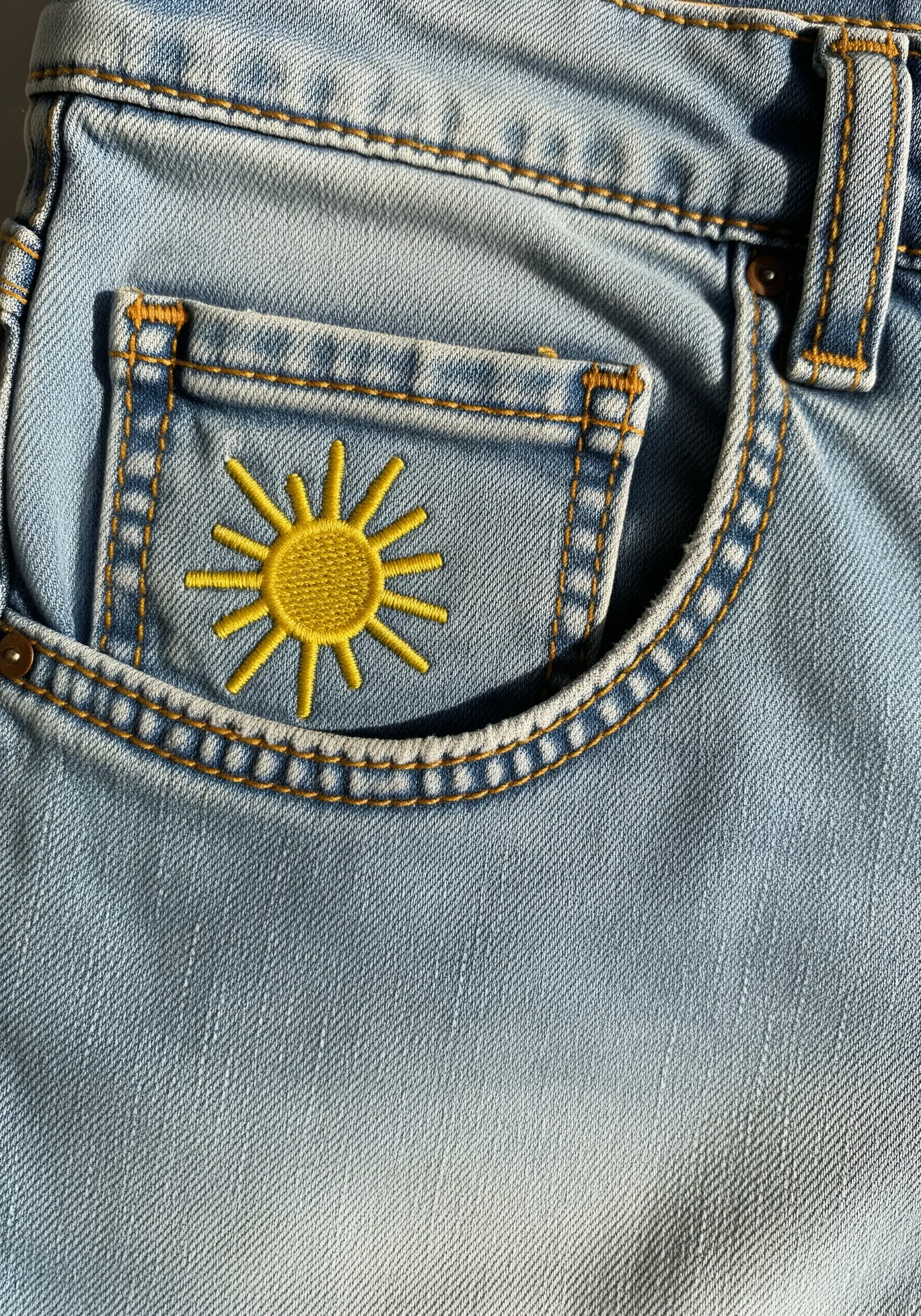 A small, bright yellow sun embroidered on the coin pocket of blue jeans.