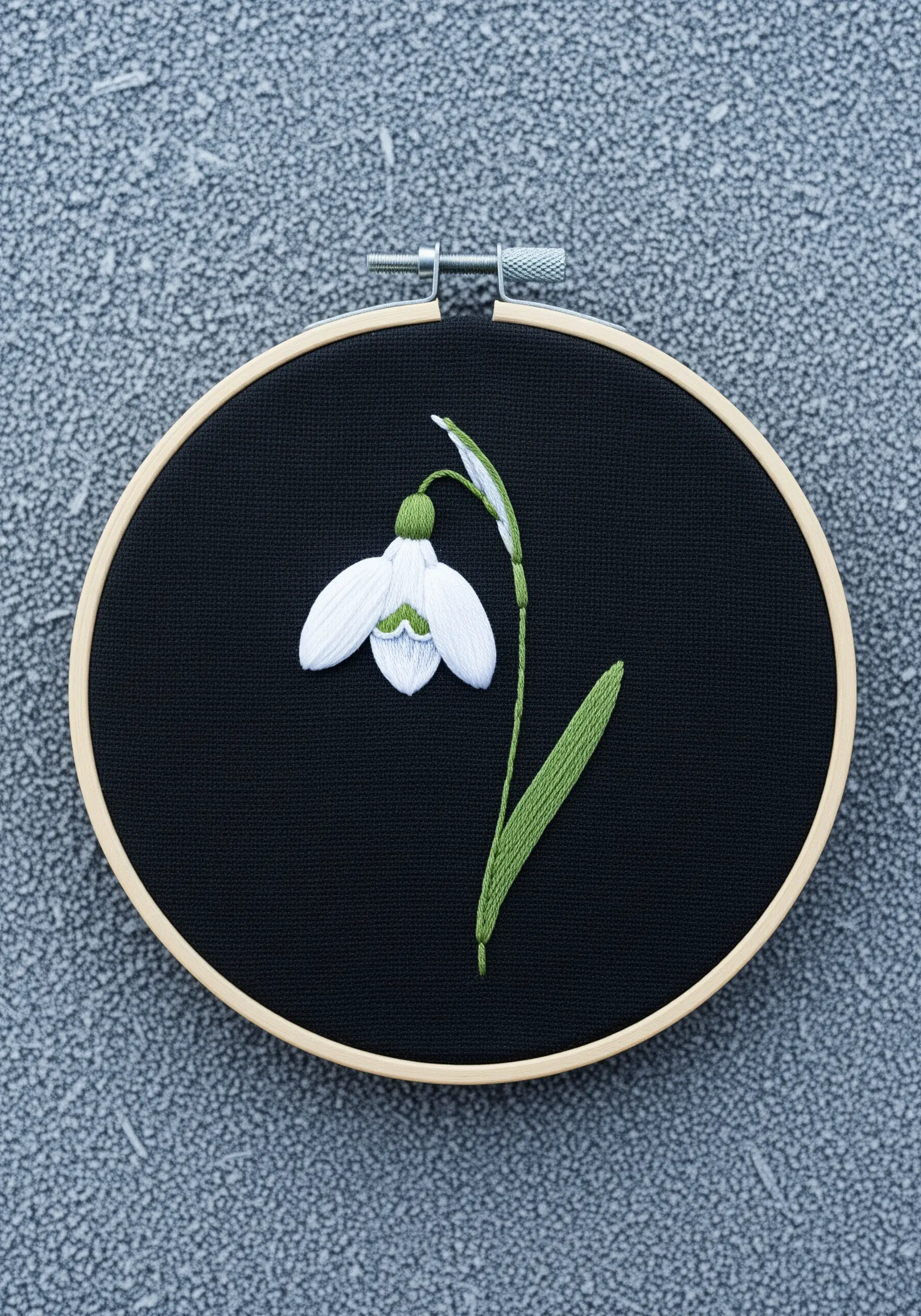 A single white snowdrop flower embroidered on black fabric inside a wooden hoop.