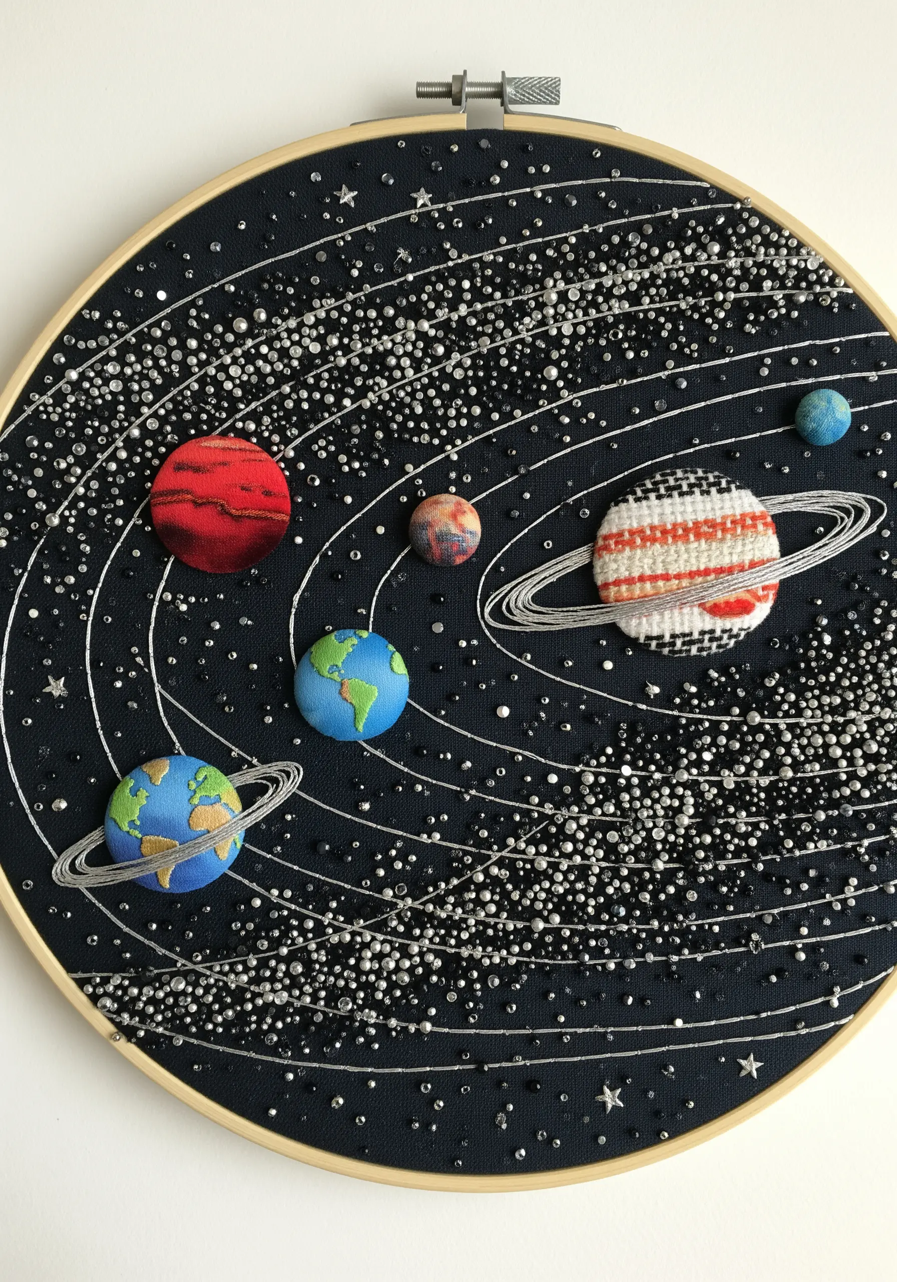 A solar system in an embroidery hoop, with woven planets and beaded stars on black fabric.